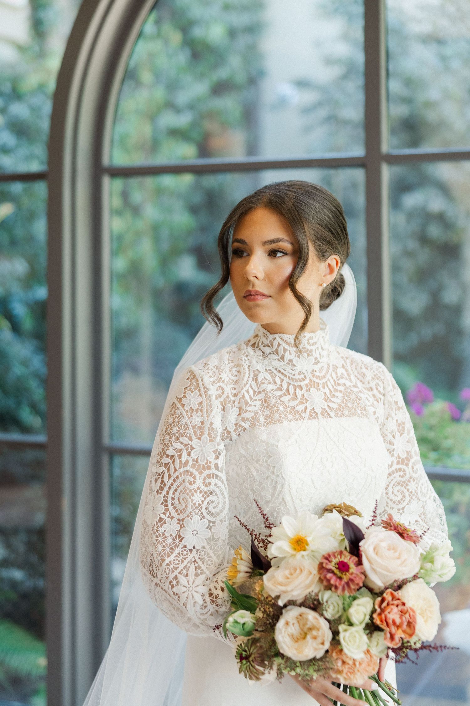 A bride in a high-neck lace wedding gown holds a romantic bouquet of peach and white flowers by an arched window.