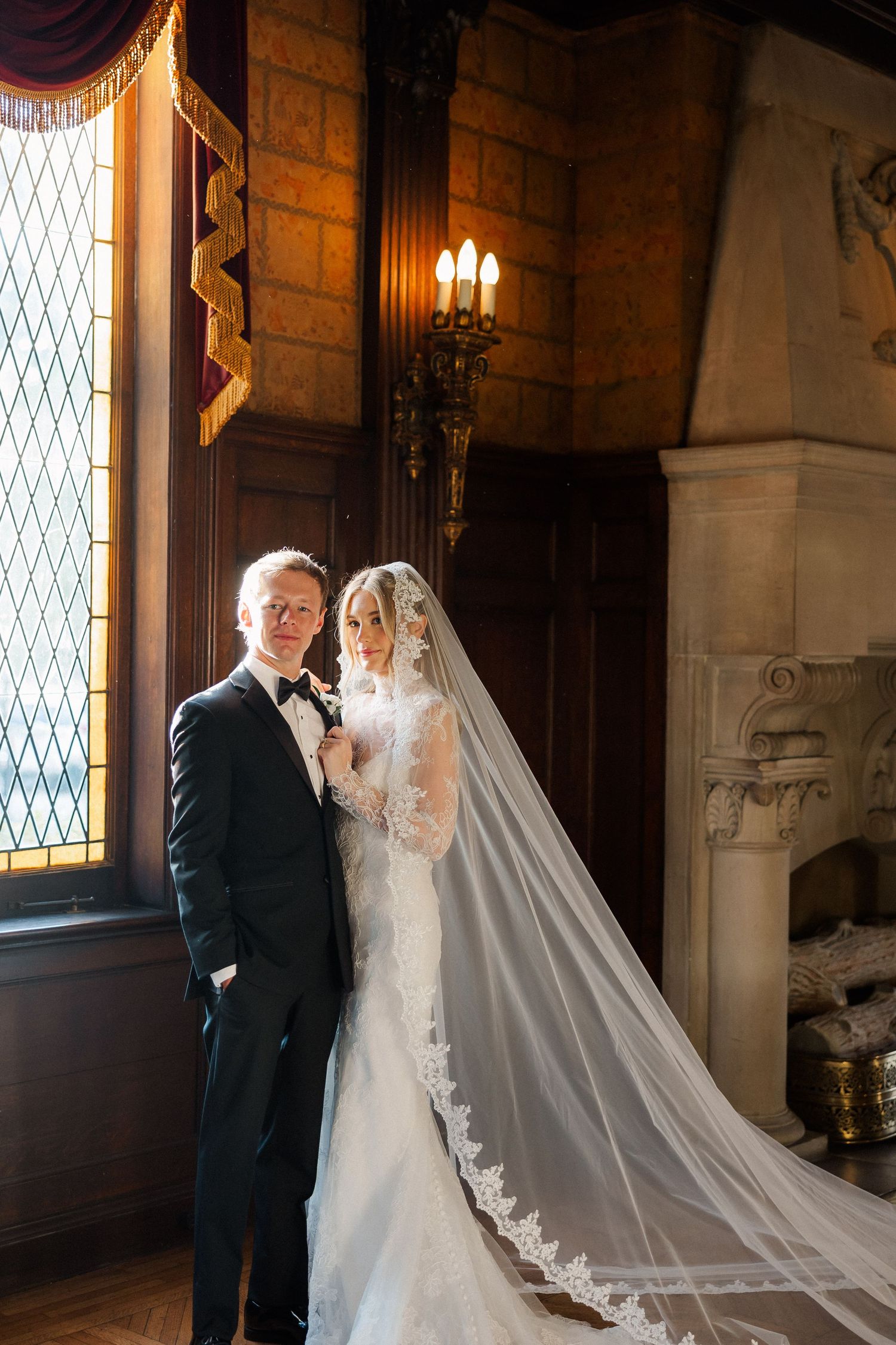 Elegant wedding couple poses in a grand historic venue with ornate windows and warm lighting.