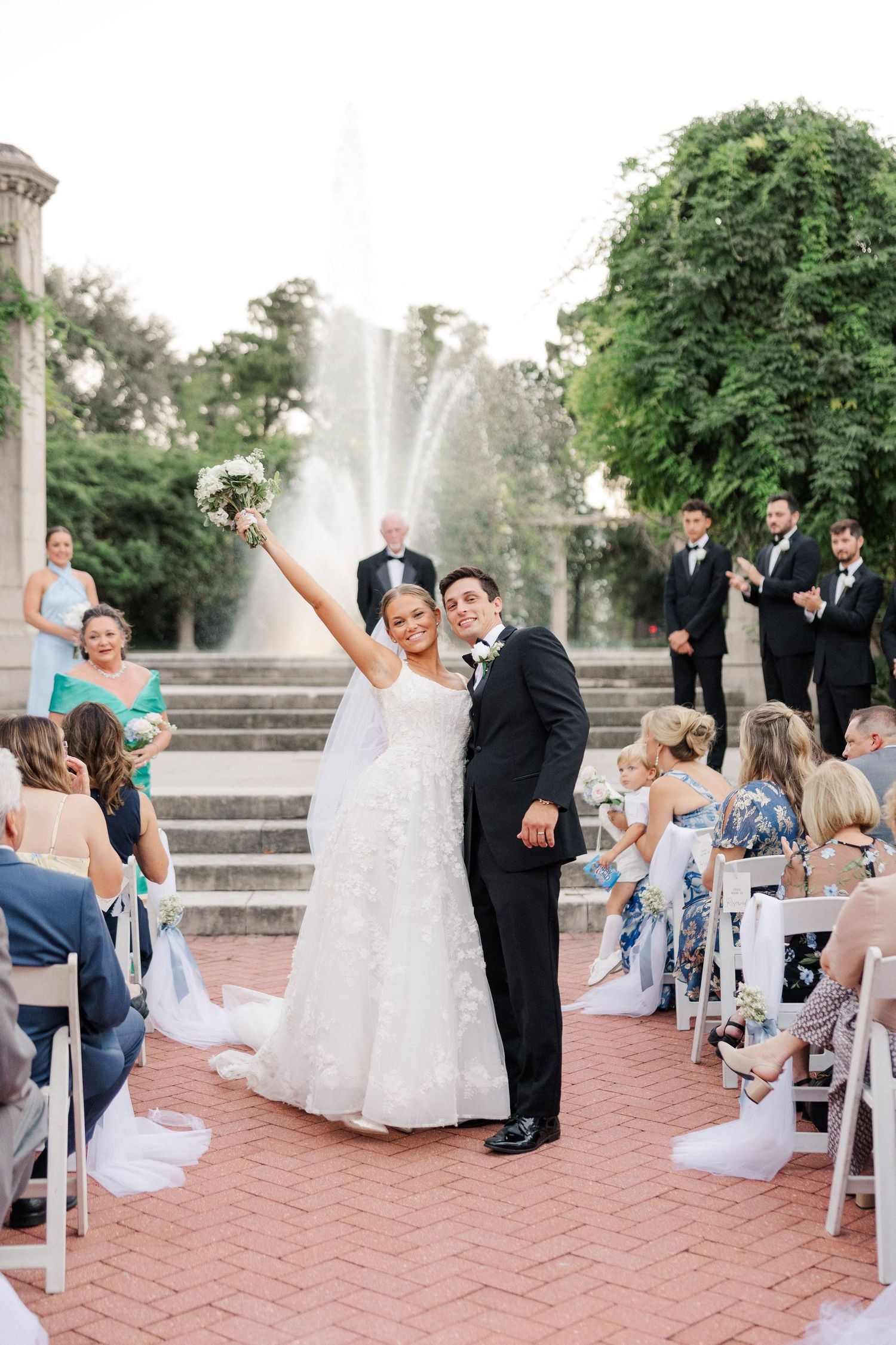 Newlyweds celebrate with raised arms at outdoor ceremony with fountain and guests in background.