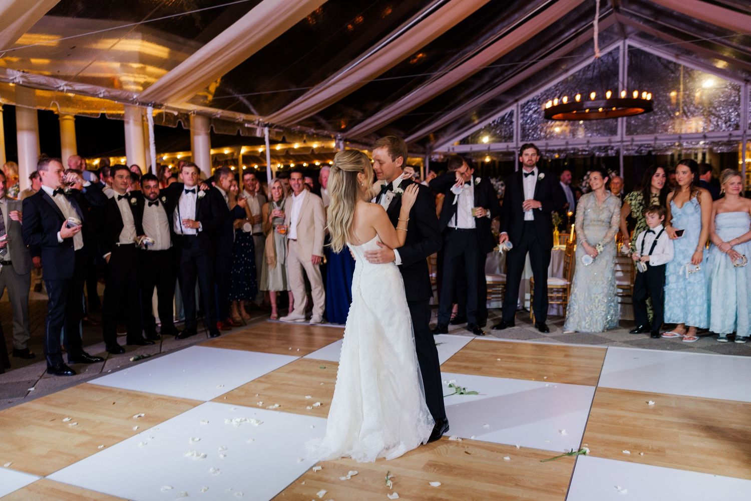 Wedding couple's first dance on checkered dance floor surrounded by guests under tent lighting.