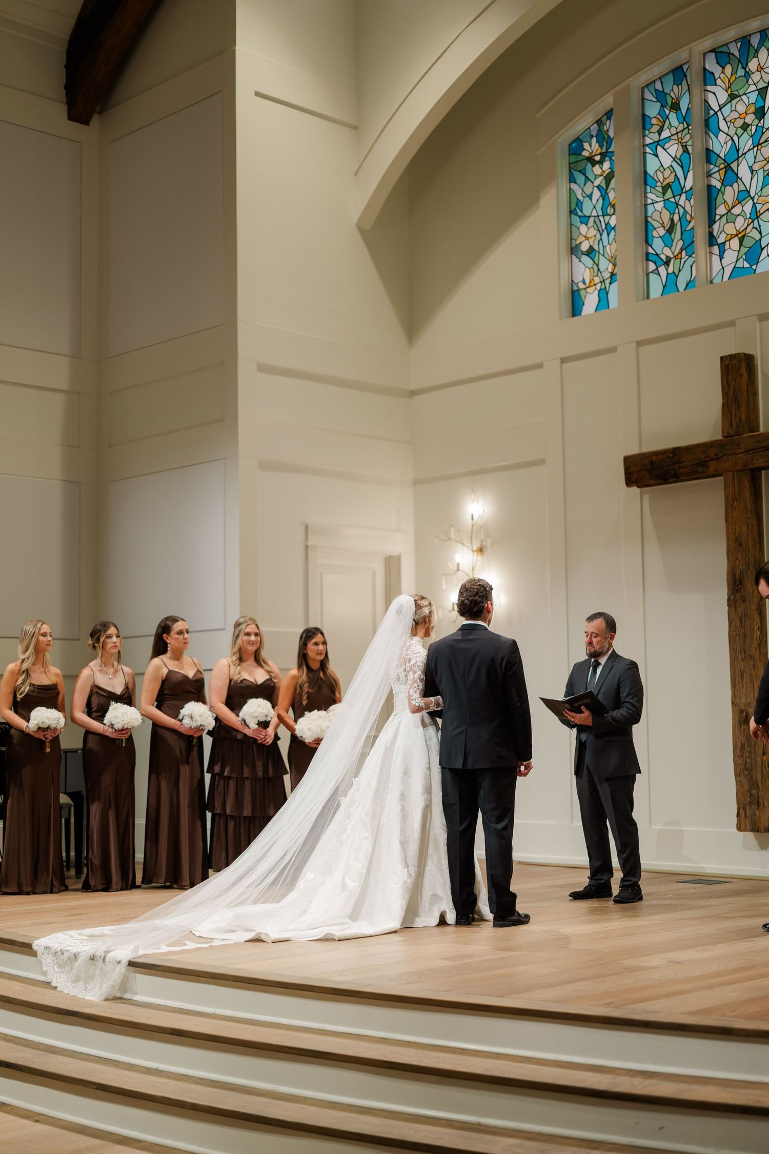 Wedding party lined up on a church stage with bridesmaids in dark dresses and stained glass windows in background.