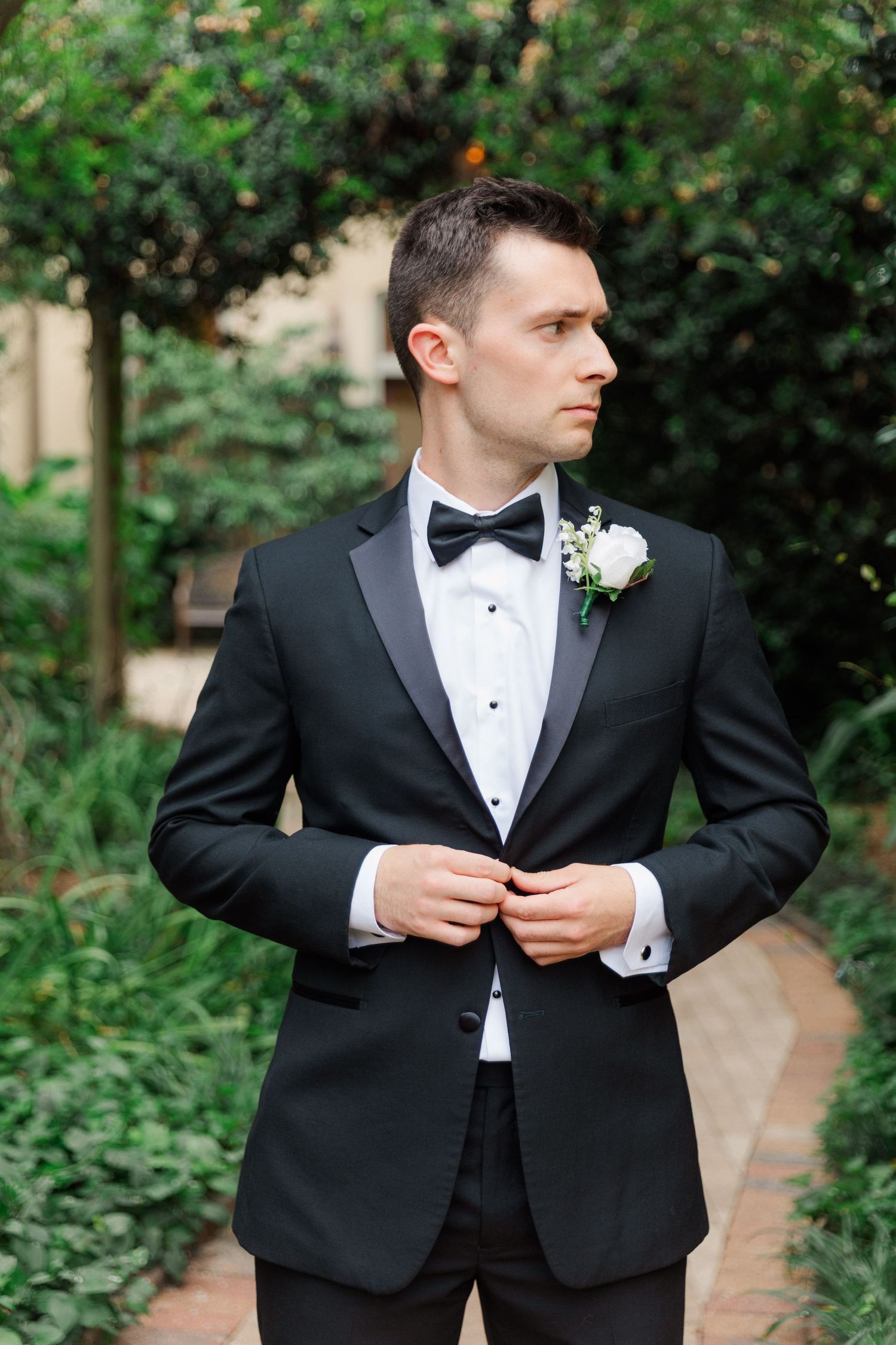 A groom in a classic black tuxedo and bowtie adjusts his jacket in a garden setting.