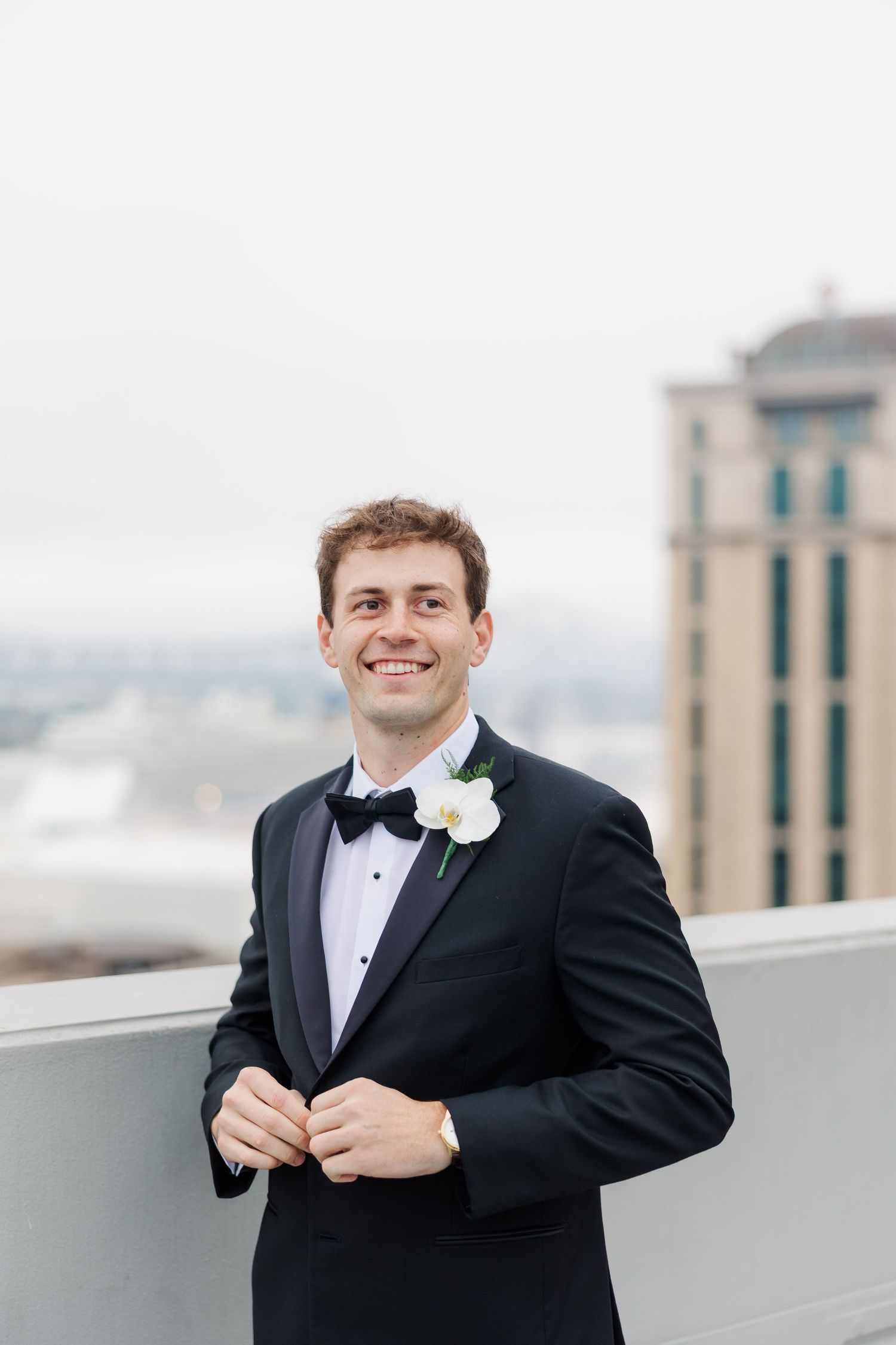 Groom in black tuxedo adjusting his bowtie on rooftop with city buildings in background.