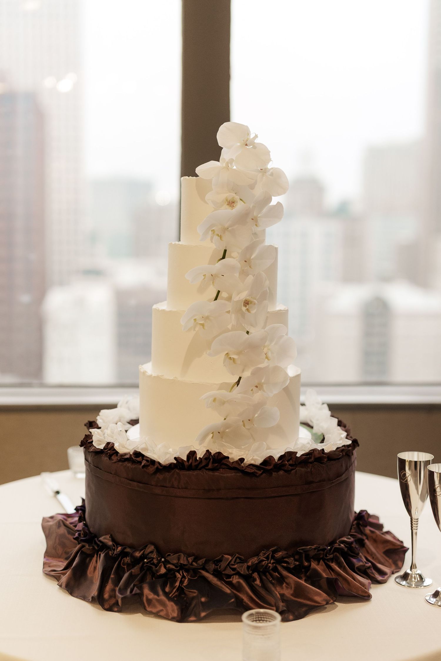 White tiered wedding cake decorated with cascading sugar flowers and chocolate ruffles at the base.