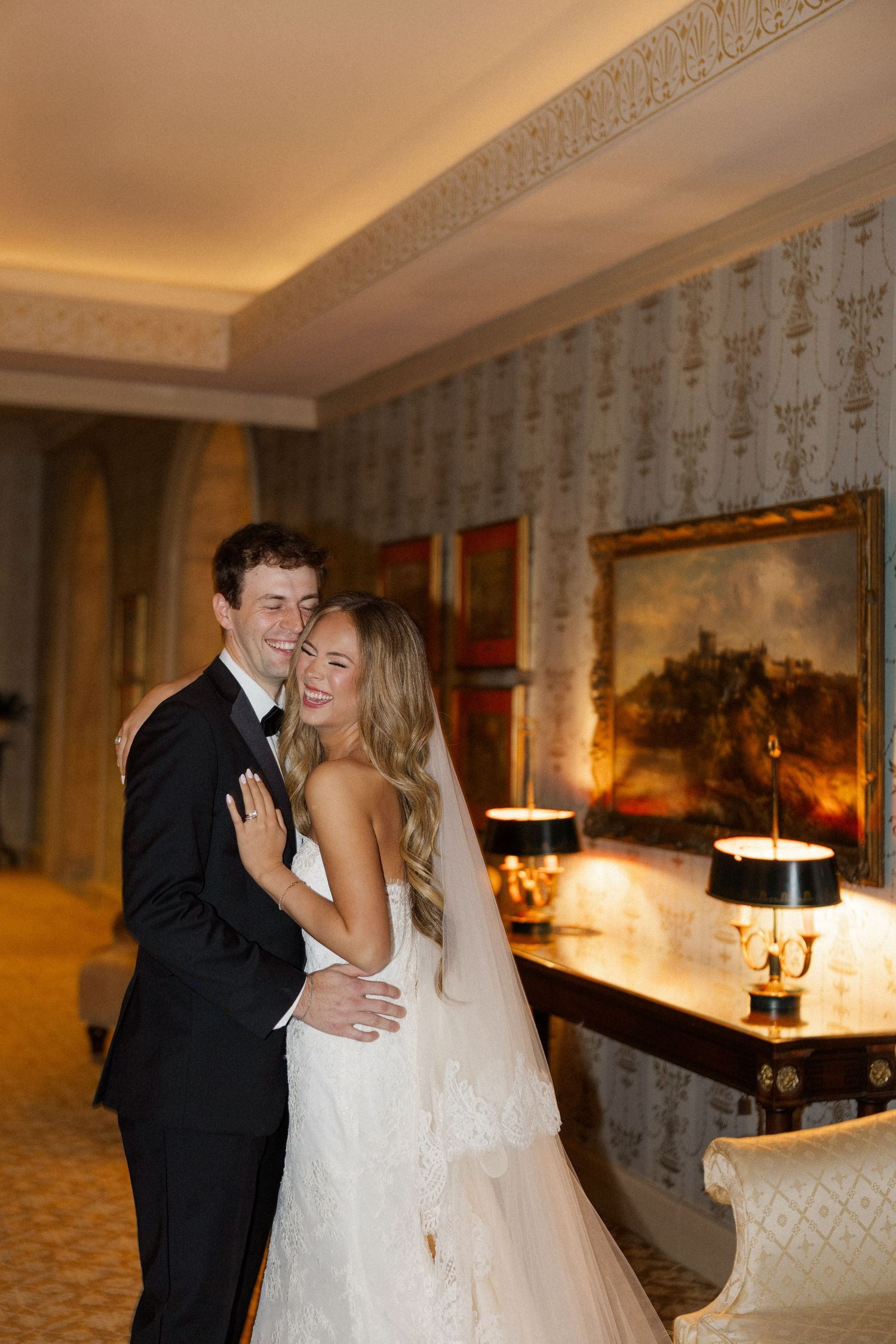 A joyful wedding couple embraces and laughs in an elegant room with vintage decor.