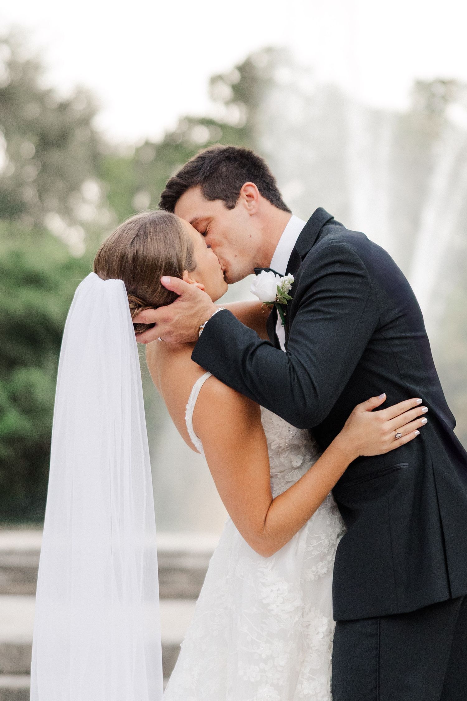 Bride and groom share a romantic first kiss in front of a fountain, bride wearing lace gown and veil.