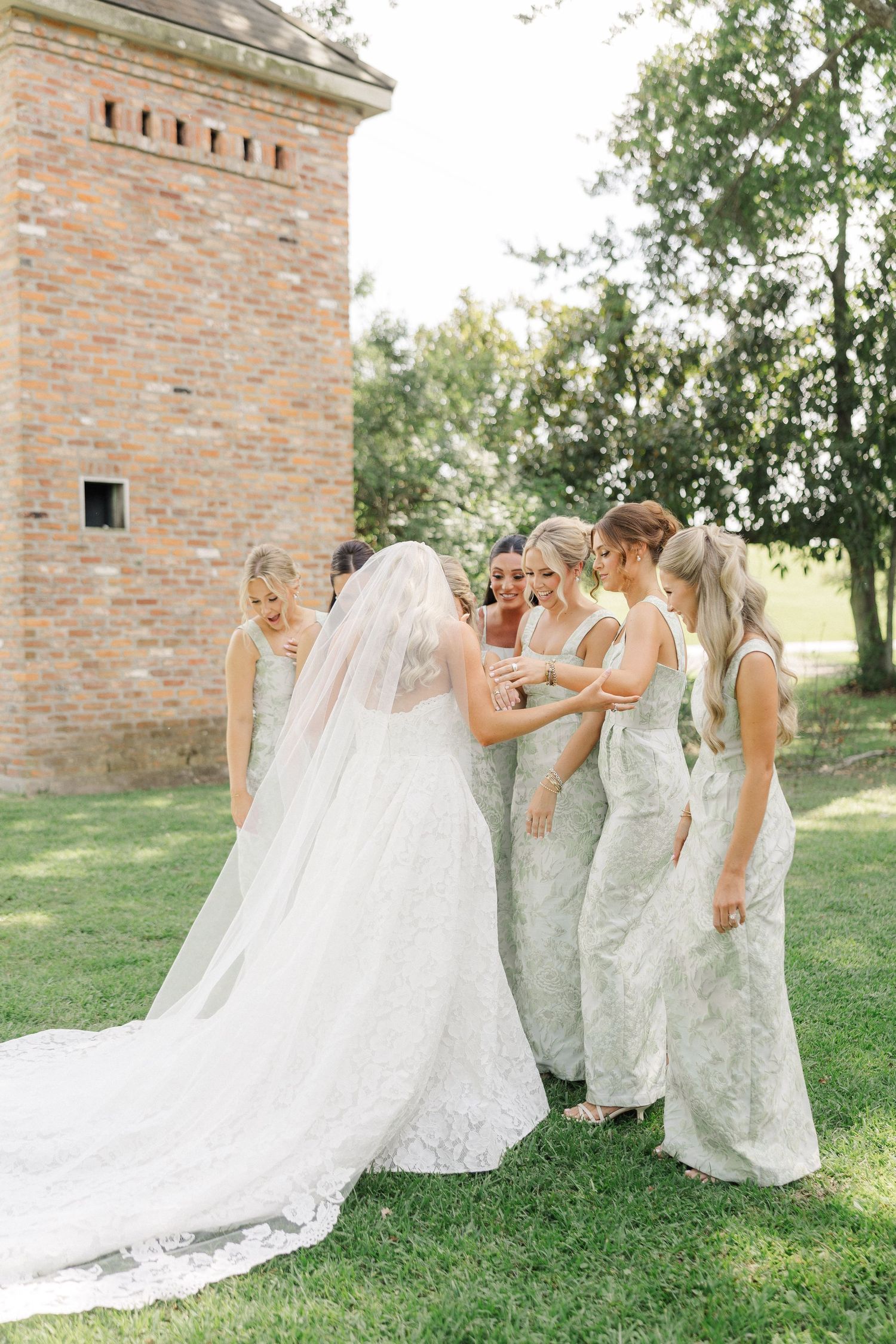Bride in lace gown with veil shares a moment with bridesmaids in sage green dresses outdoors near brick tower.