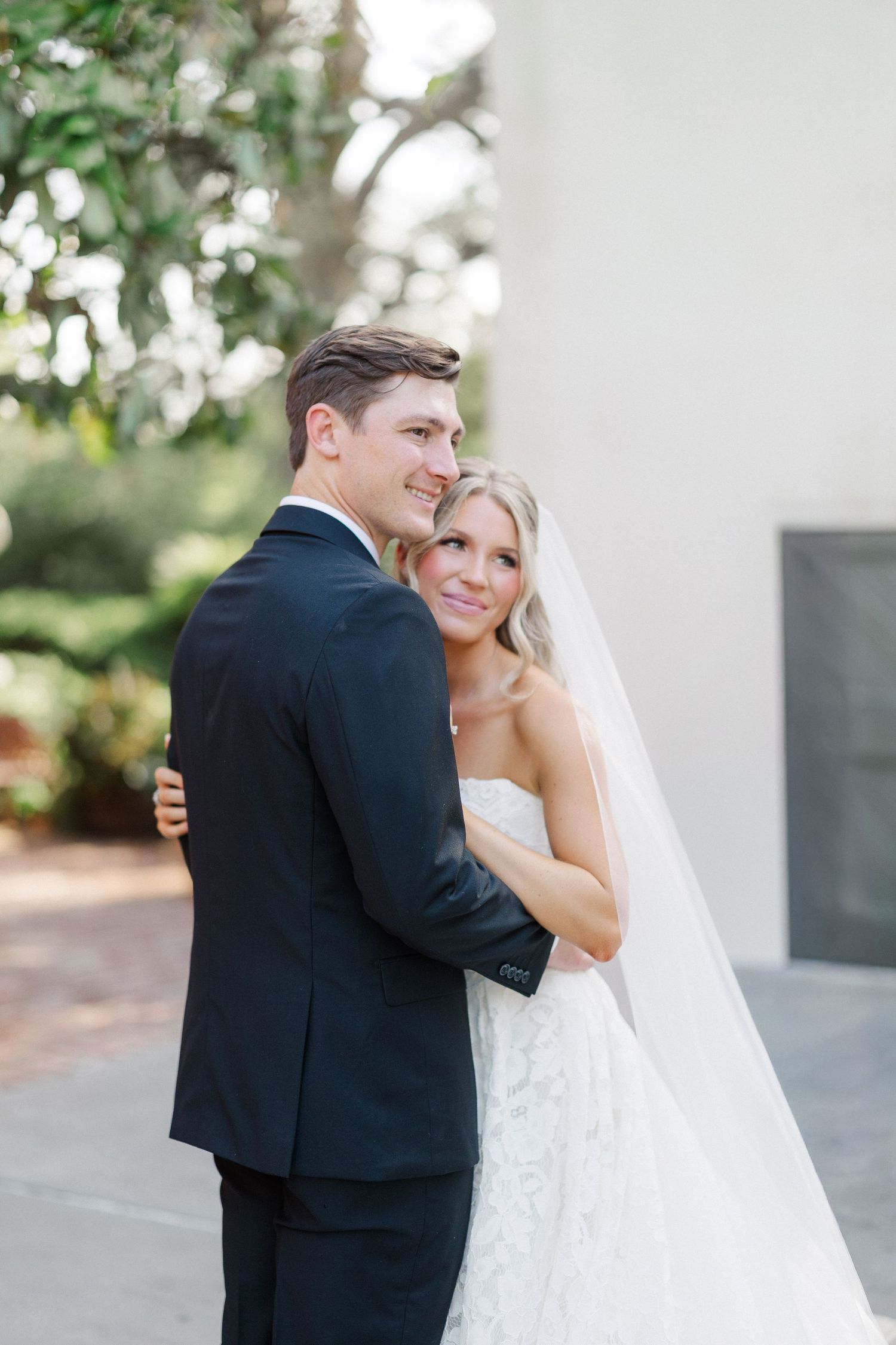 Bride in lace gown and veil embraces groom in navy suit outdoors at elegant wedding venue.
