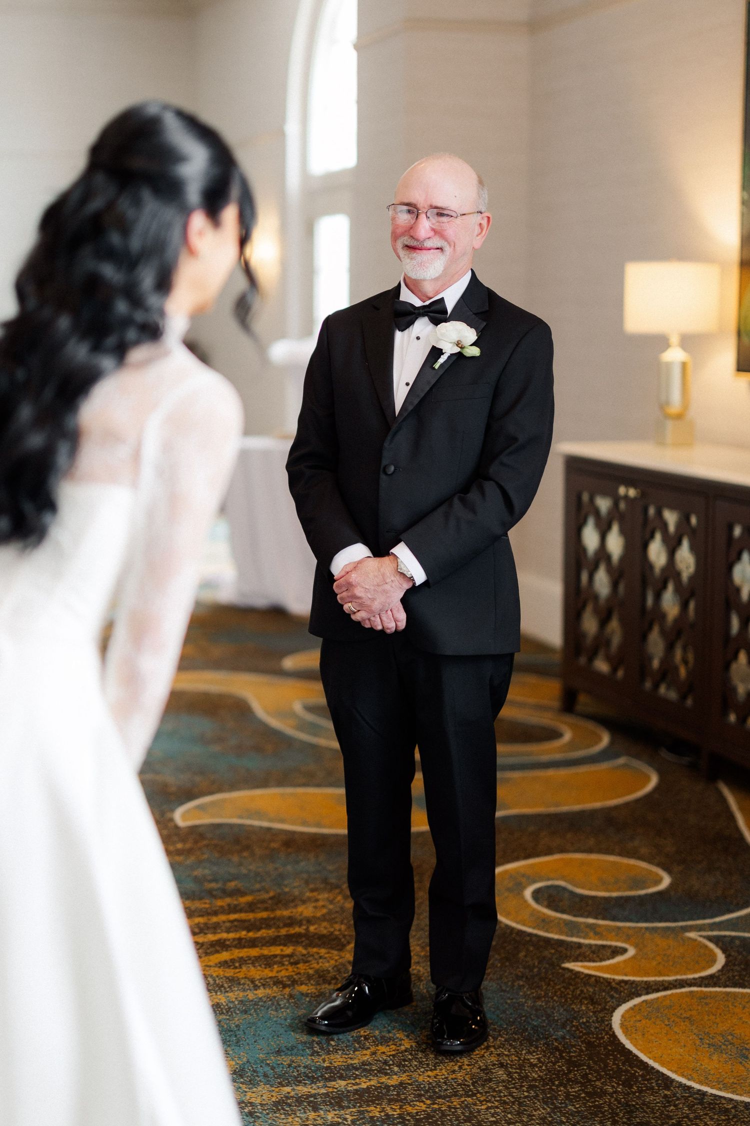 Father smiles with clasped hands seeing bride in white wedding dress during emotional first look moment.