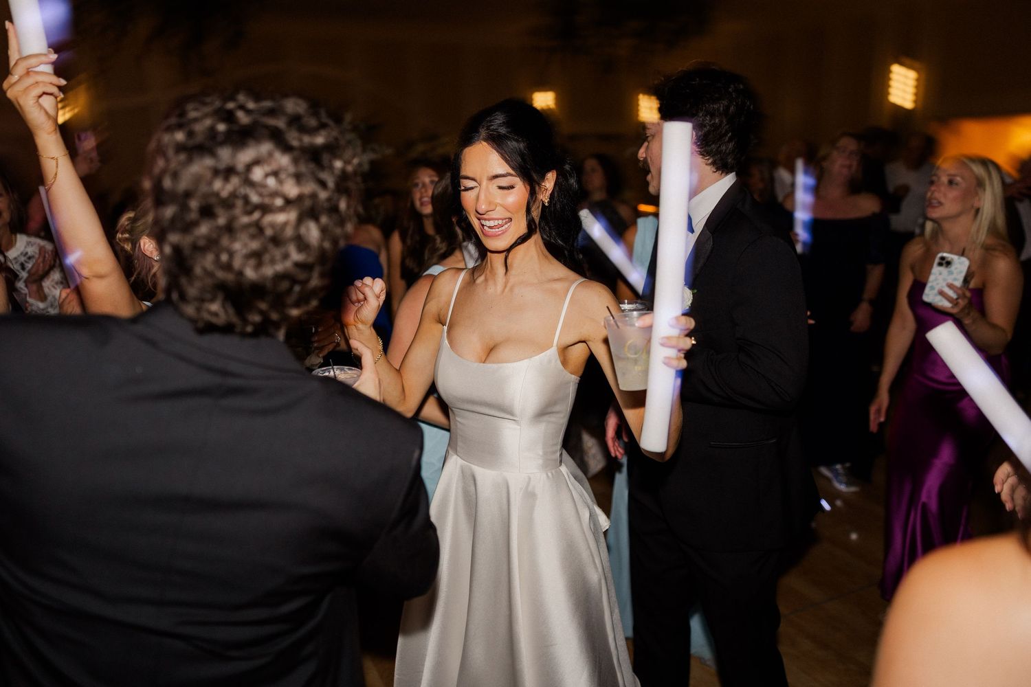 Bride in silver satin gown dancing joyfully with guests holding glow sticks at elegant wedding reception.