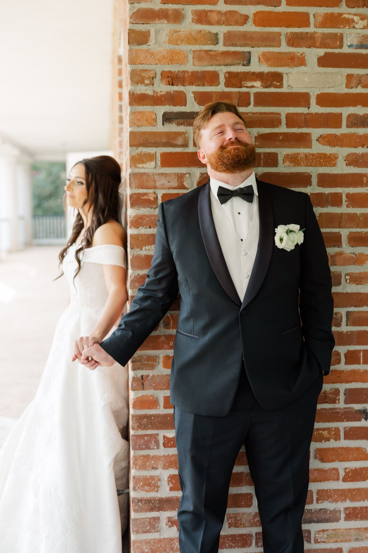 Wedding party members in formal attire pose against rustic brick wall during photo session.