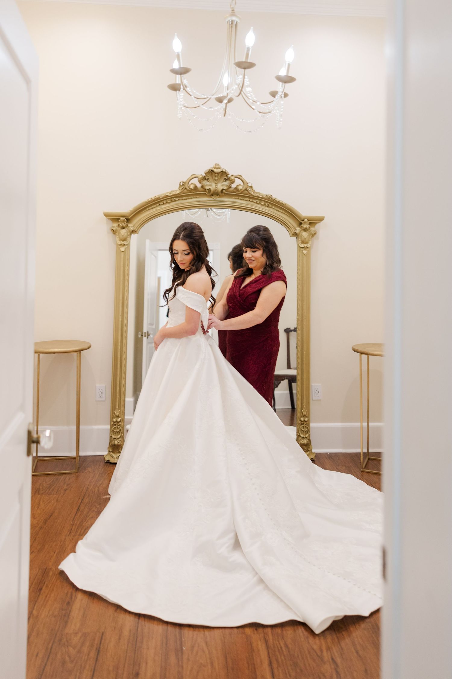 Bride's flowing white wedding gown is adjusted in elegant dressing room with ornate gold mirrors.