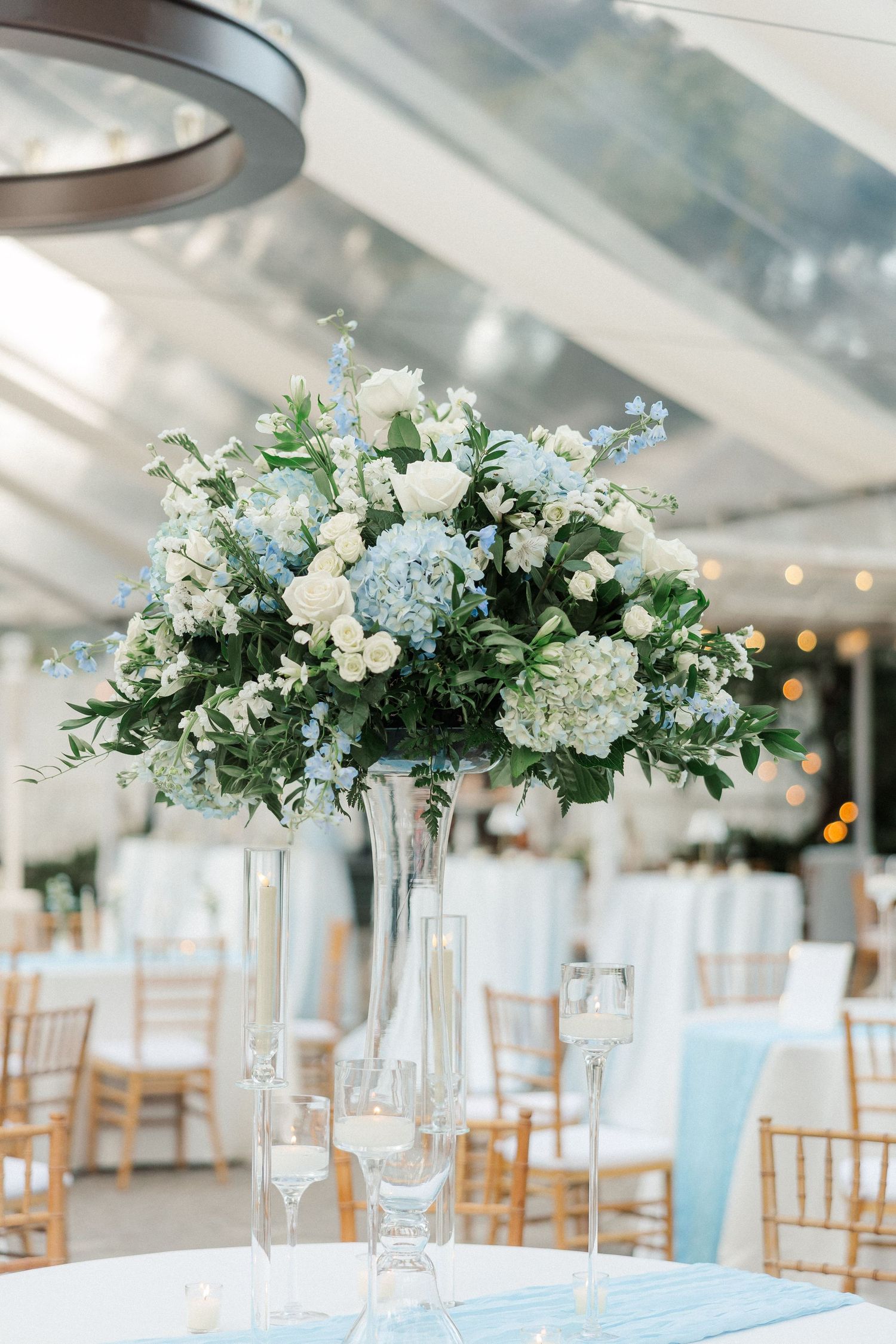 Elegant white and light blue floral centerpieces in tall glass vases decorate tables at a wedding reception.