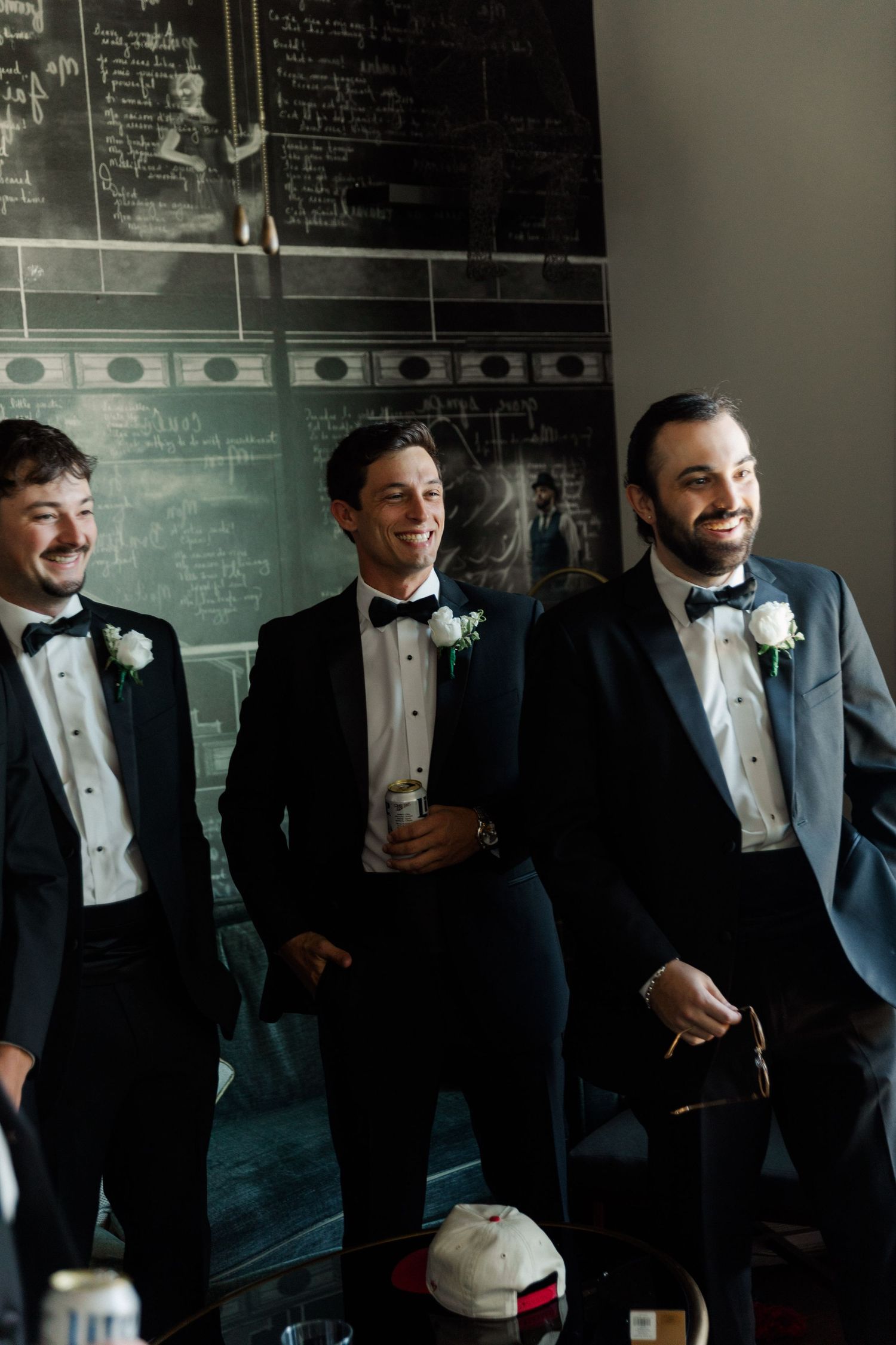 Three groomsmen in tuxedos share a candid moment laughing together in front of a chalkboard wall.