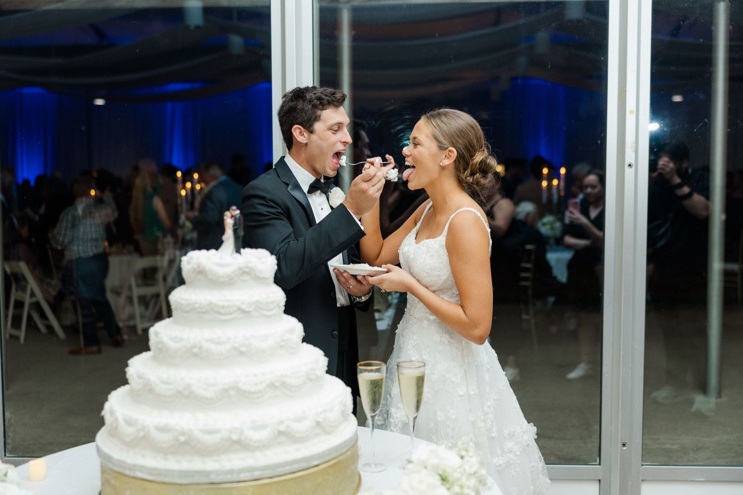 A wedding couple feeds each other bites of their elegant white tiered wedding cake at the reception.
