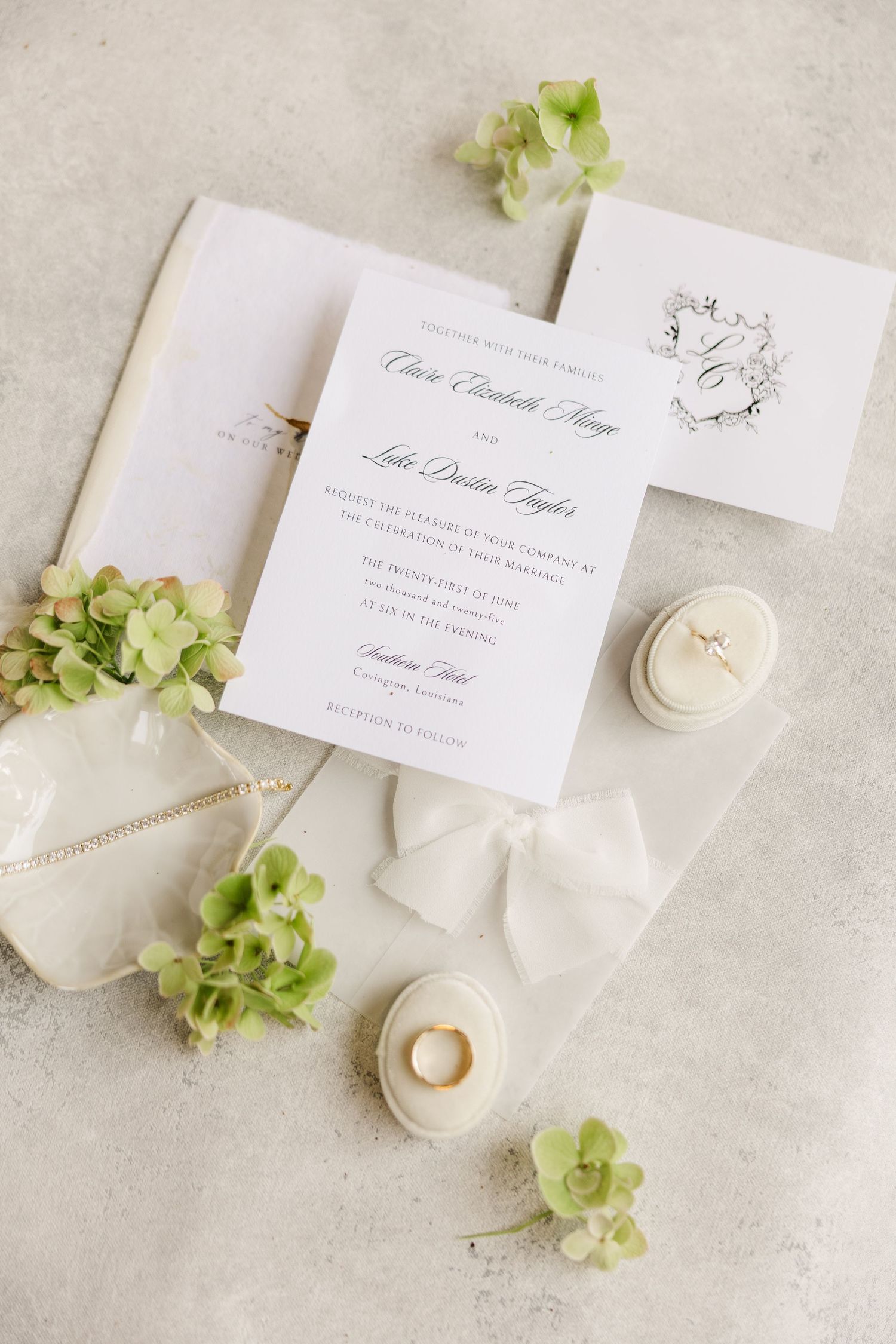 Elegant wedding invitation suite with white stationery, green hydrangeas, and delicate gold jewelry on a light textured surface.