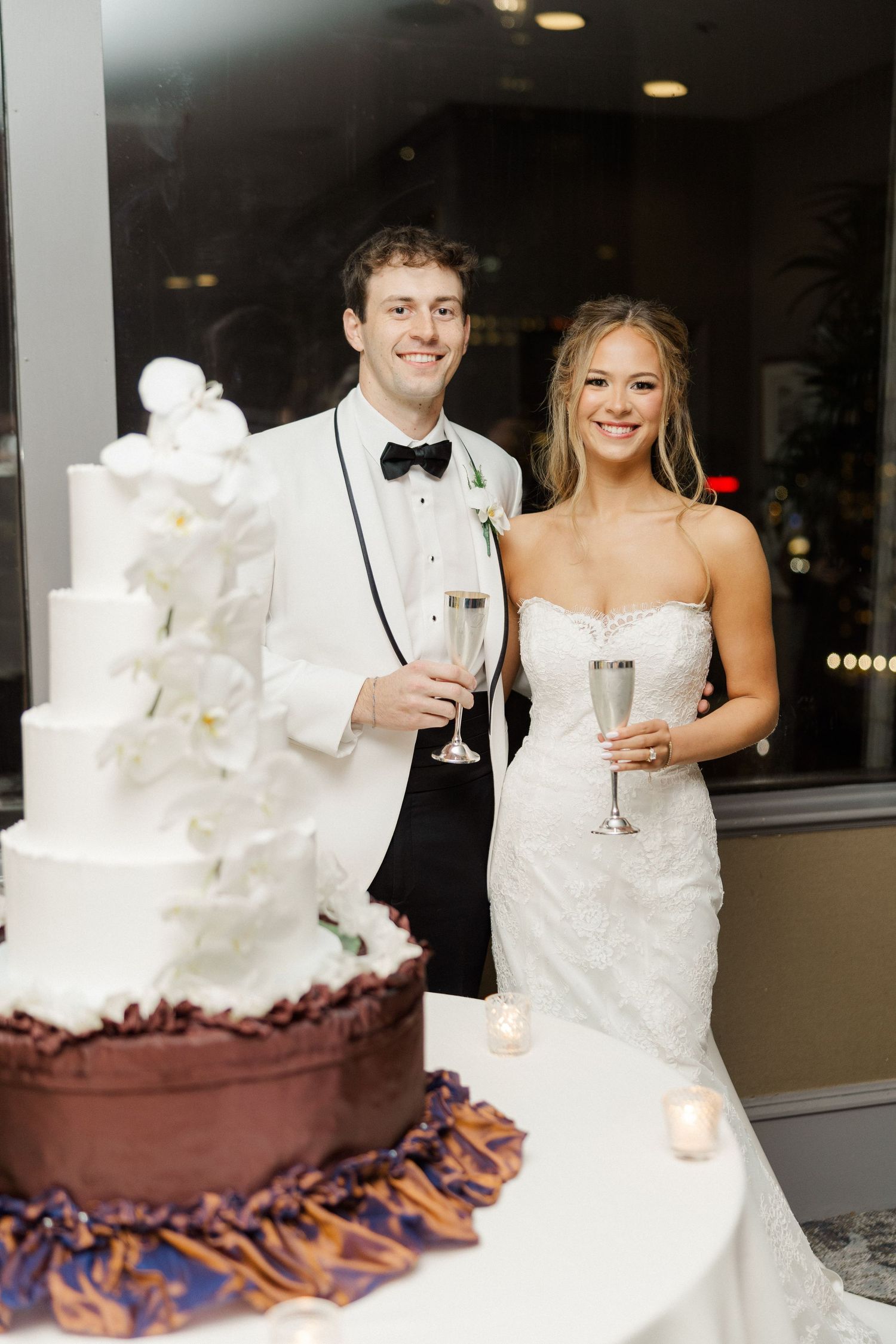 A wedding couple poses next to their elegant white and chocolate tiered wedding cake at their reception.
