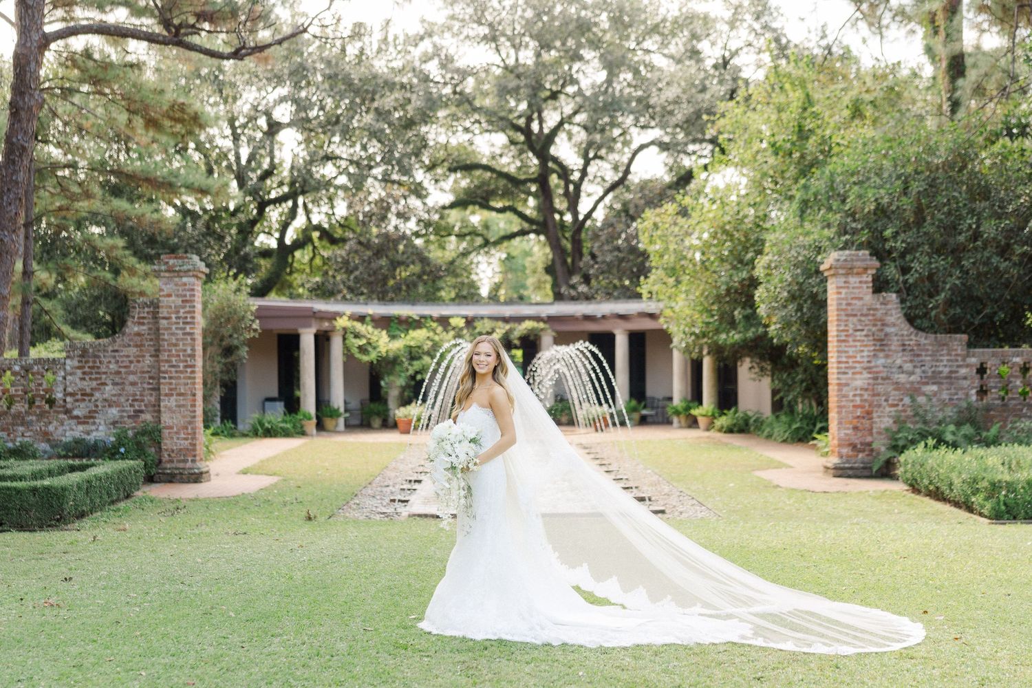 A bride in a flowing white wedding dress walks through a scenic garden path lined with trees and brick columns.