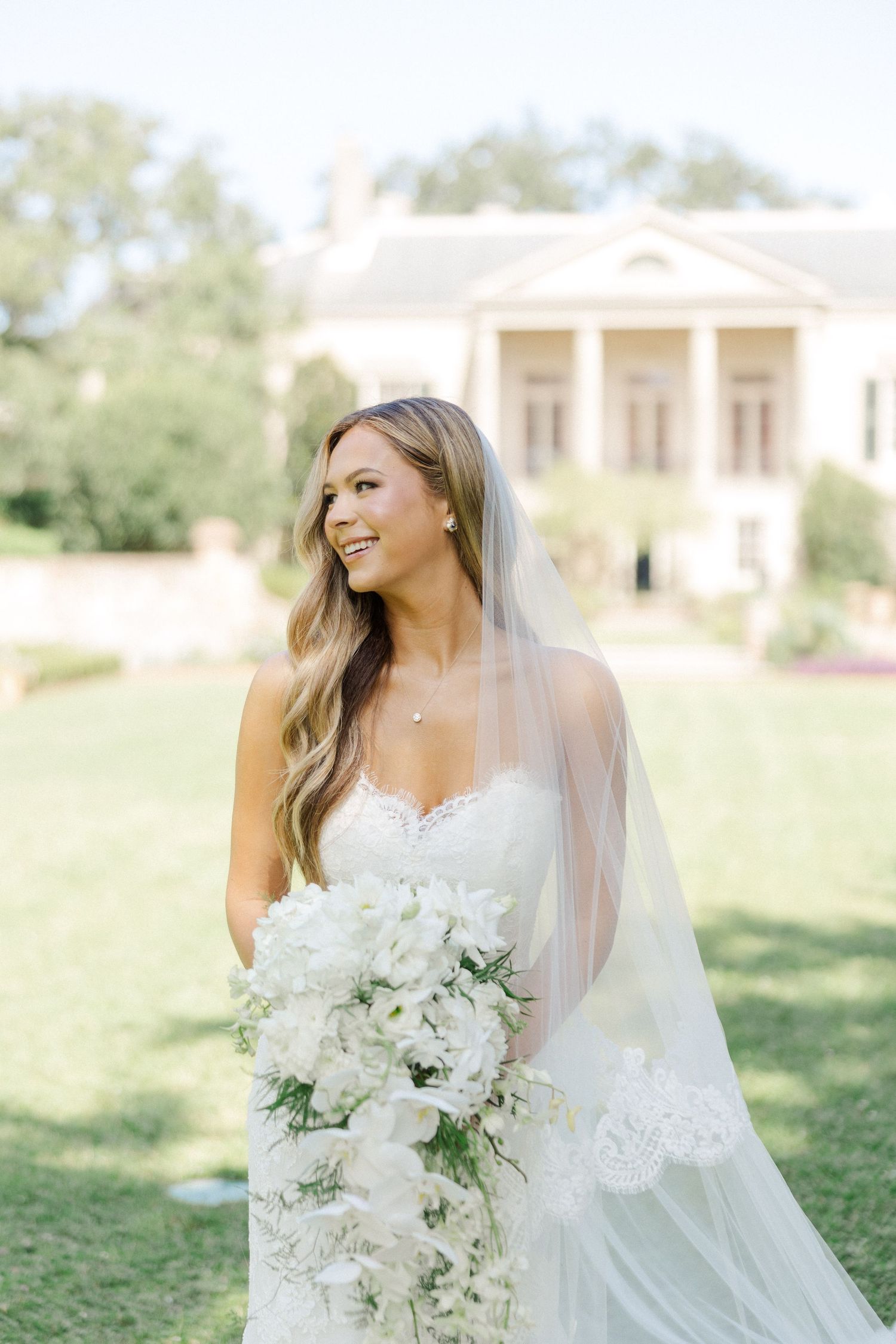 A bride in a white lace wedding dress holds an elegant white floral bouquet while standing on a sunny lawn.