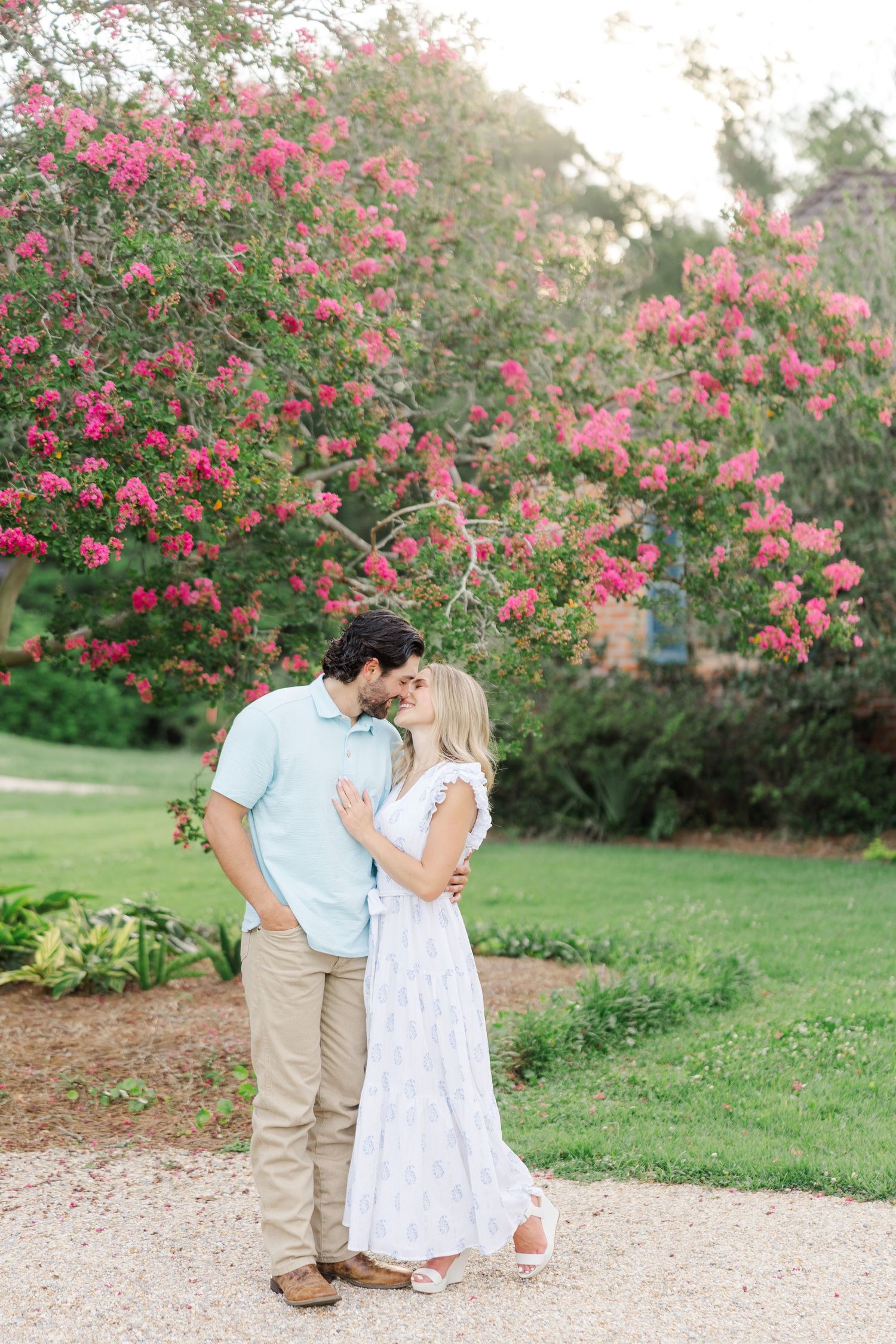 A couple shares a romantic moment beneath blooming pink crepe myrtle trees in a garden setting.