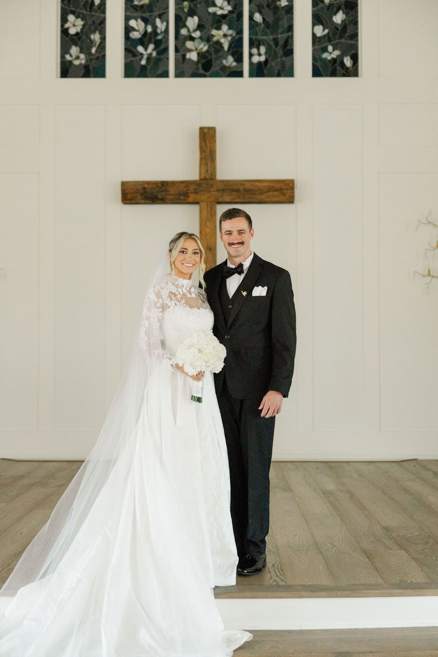 Bride and groom pose in front of white wall with decorative cross in church setting.