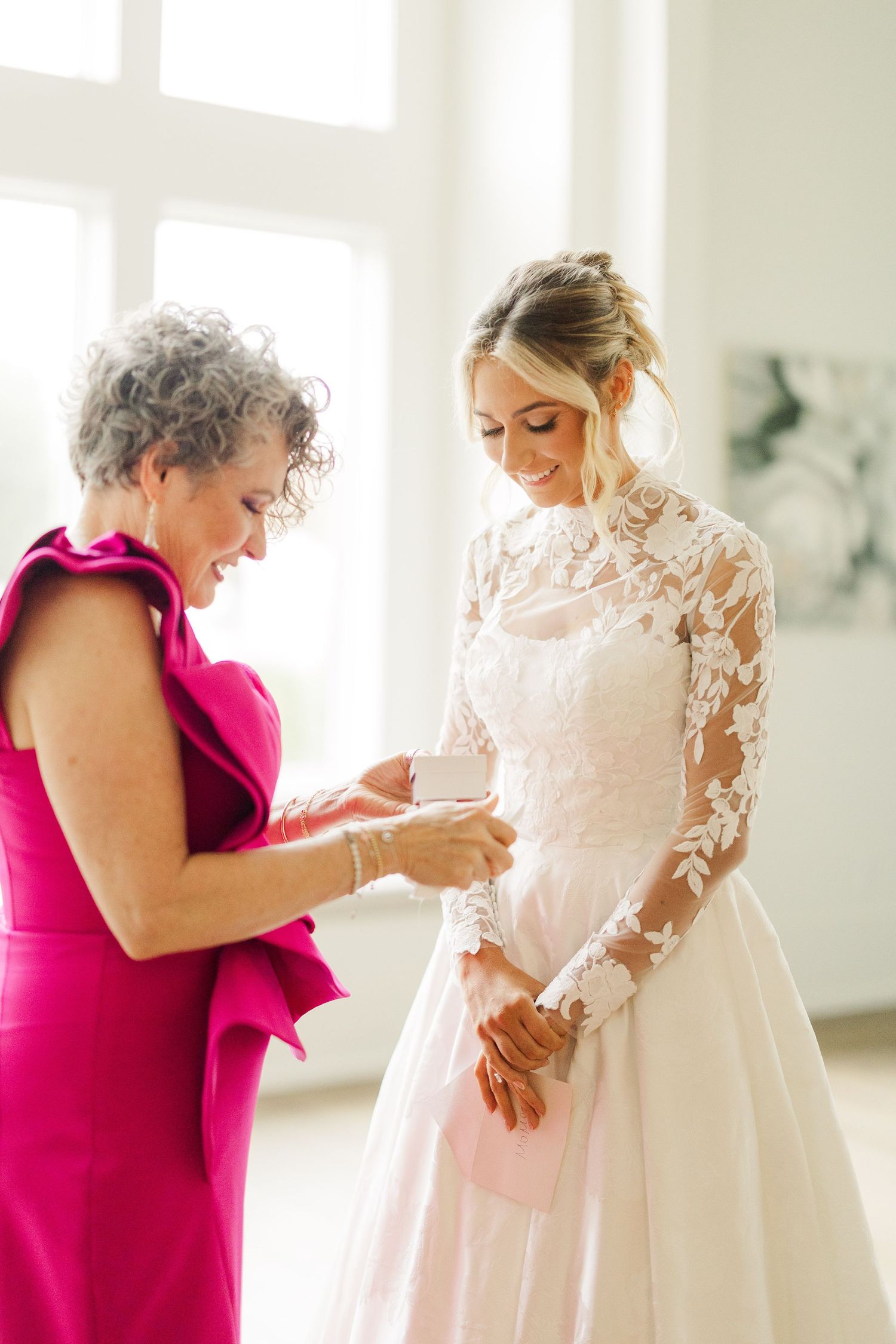A woman in a hot pink dress helps adjust a delicate lace wedding gown in a bright sunlit room.