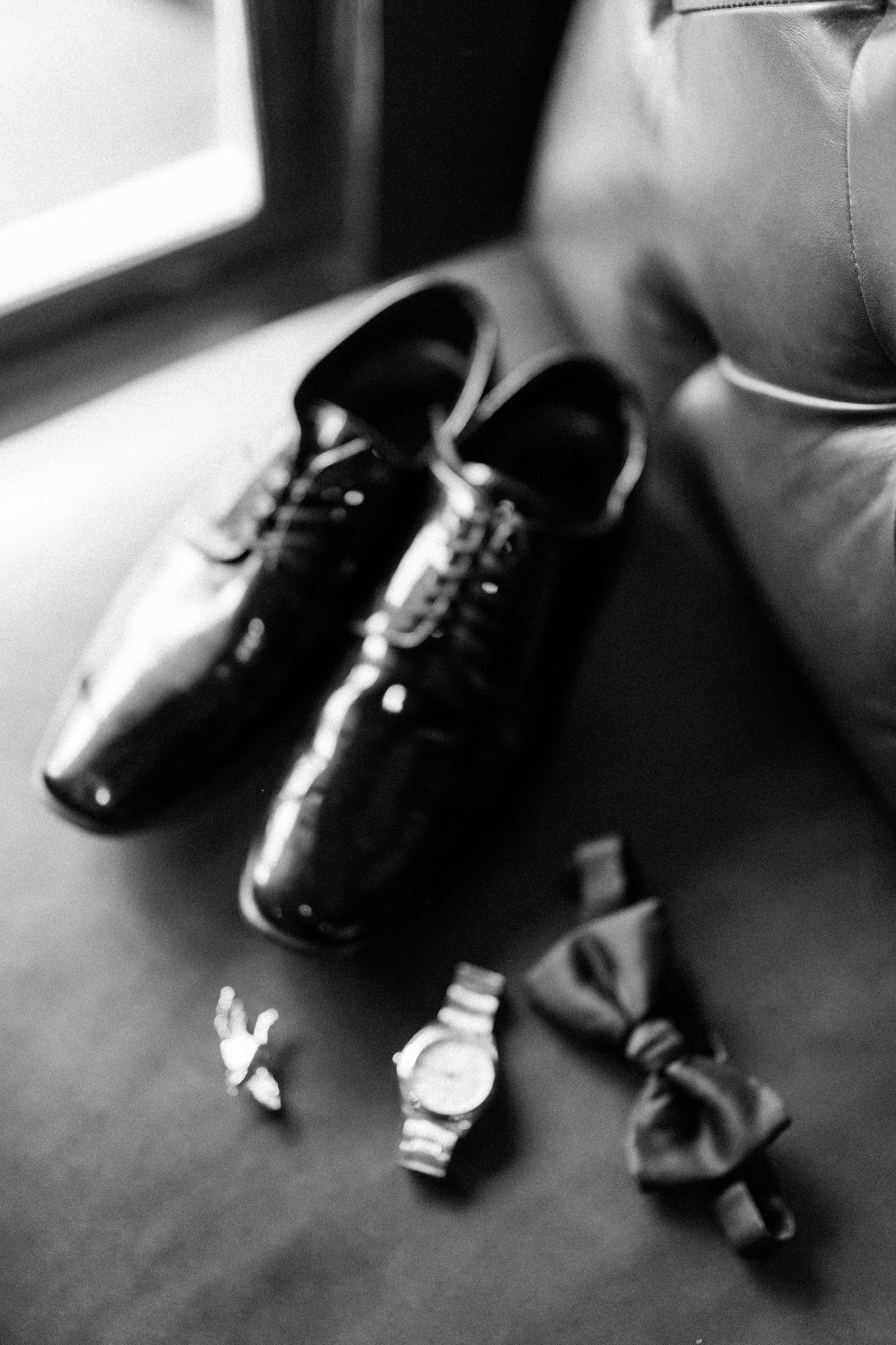 Black and white photo of formal dress shoes, bow tie, and watch arranged on dark surface near window.