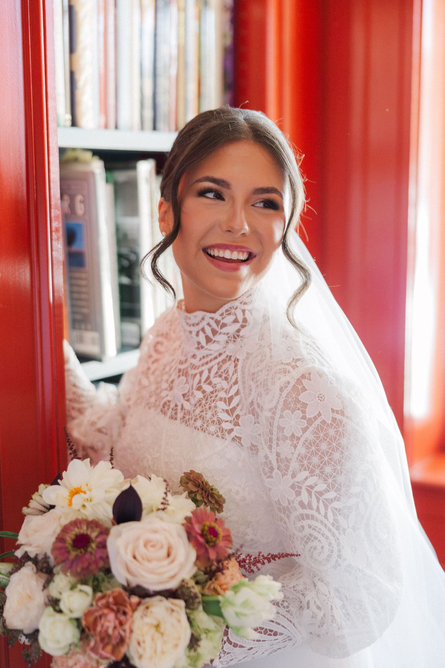 A radiant bride poses in front of vibrant red walls while holding a romantic bouquet of blush and white flowers.
