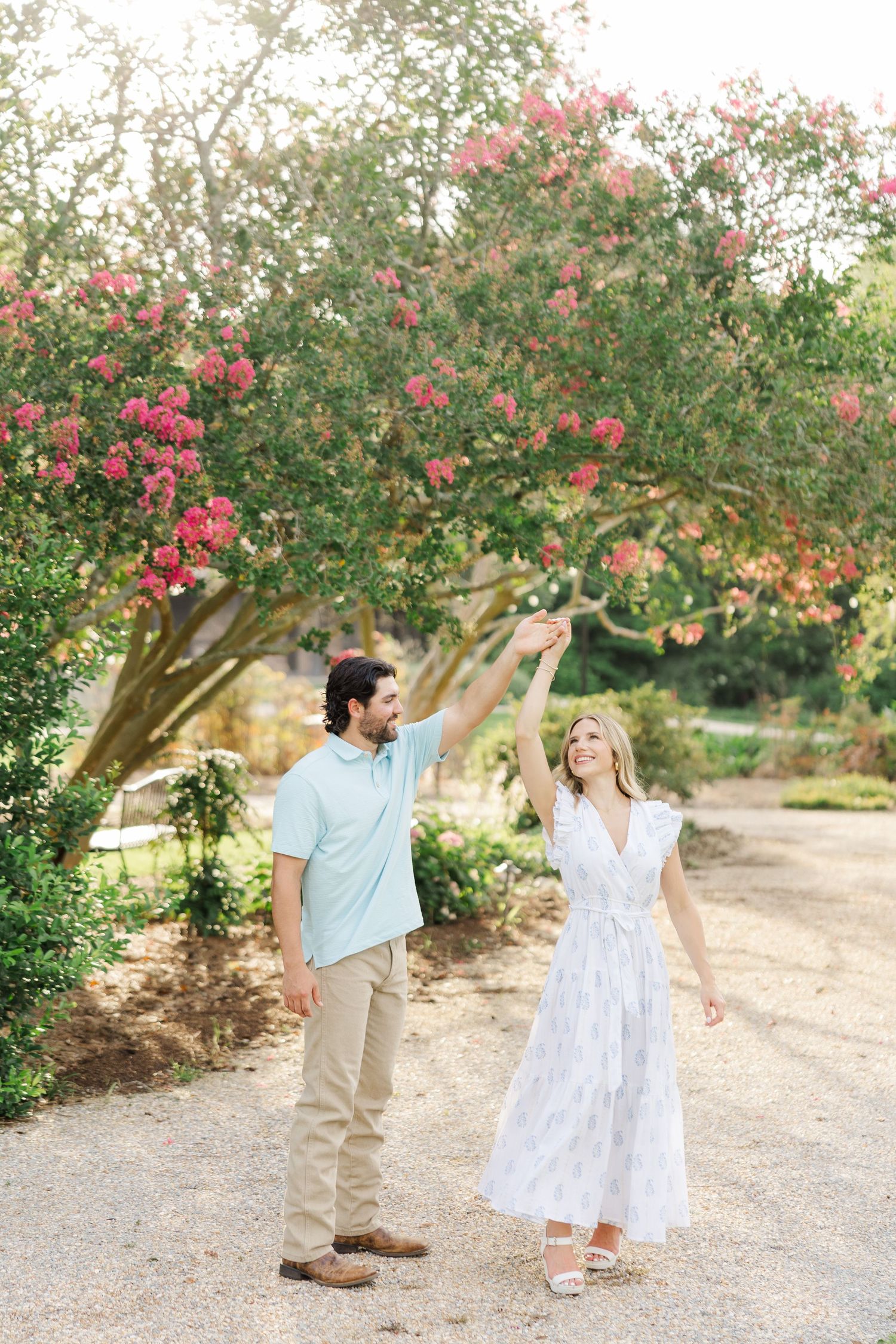 A couple dances and twirls together on a path beneath blooming pink trees in a romantic garden setting.
