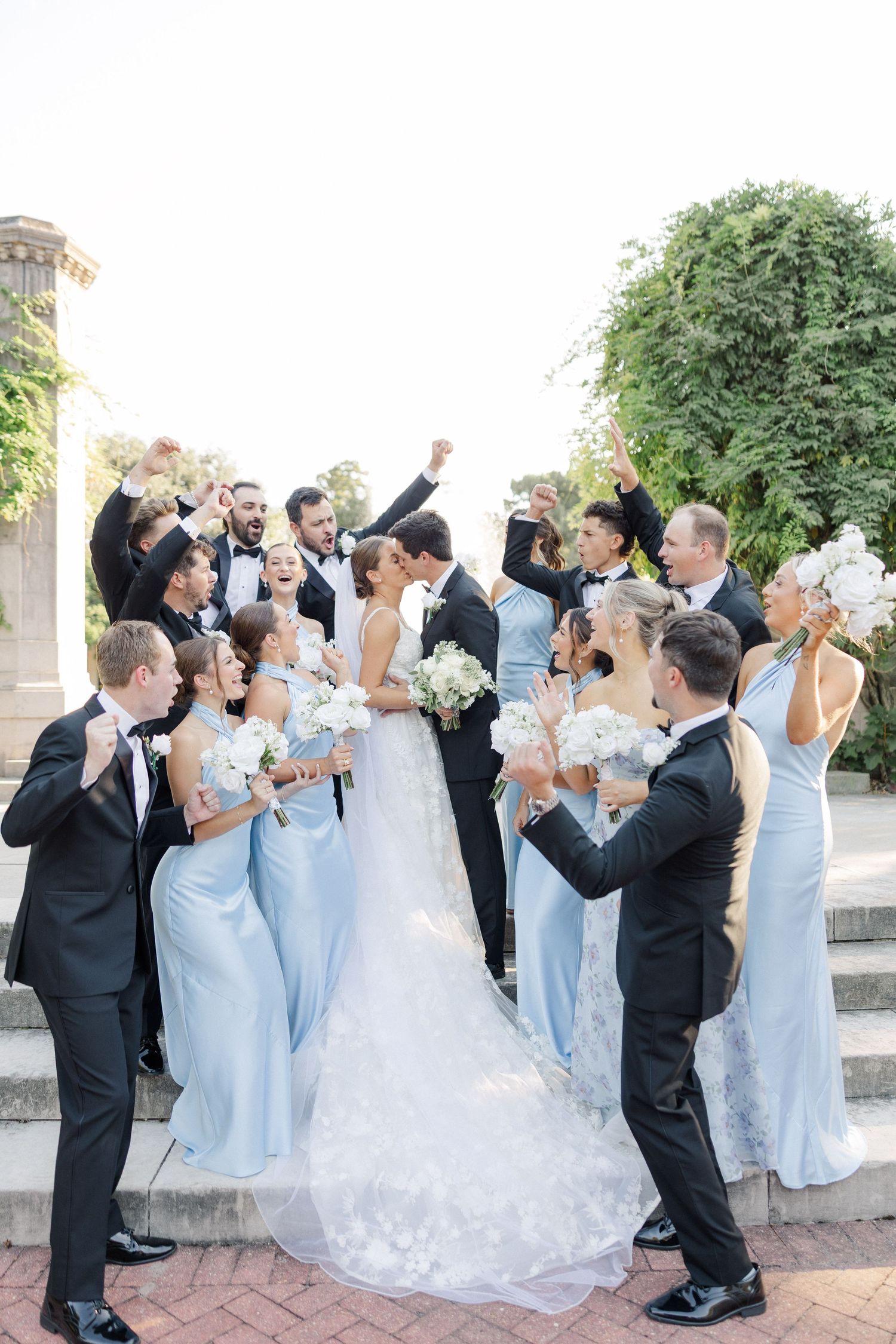 Wedding party in black tuxedos and light blue dresses raise their hands in celebration on outdoor stone steps.