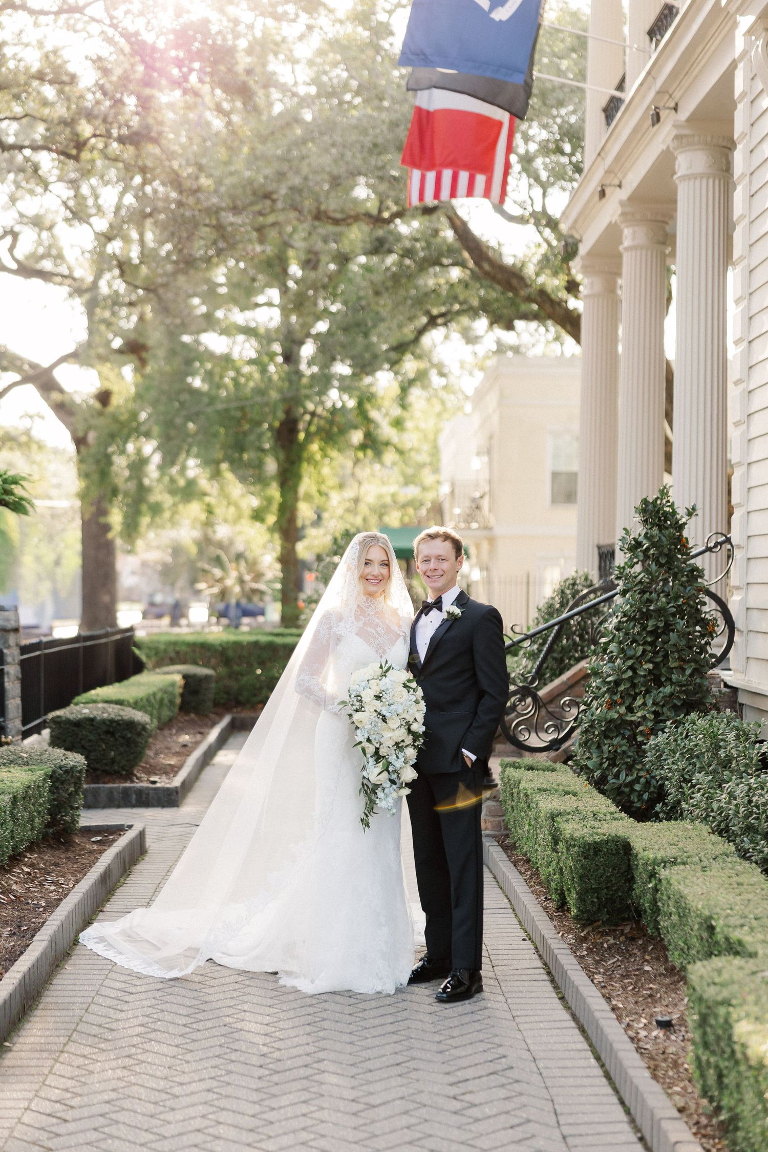 Elegant wedding couple poses along a columned walkway with American flags and greenery.