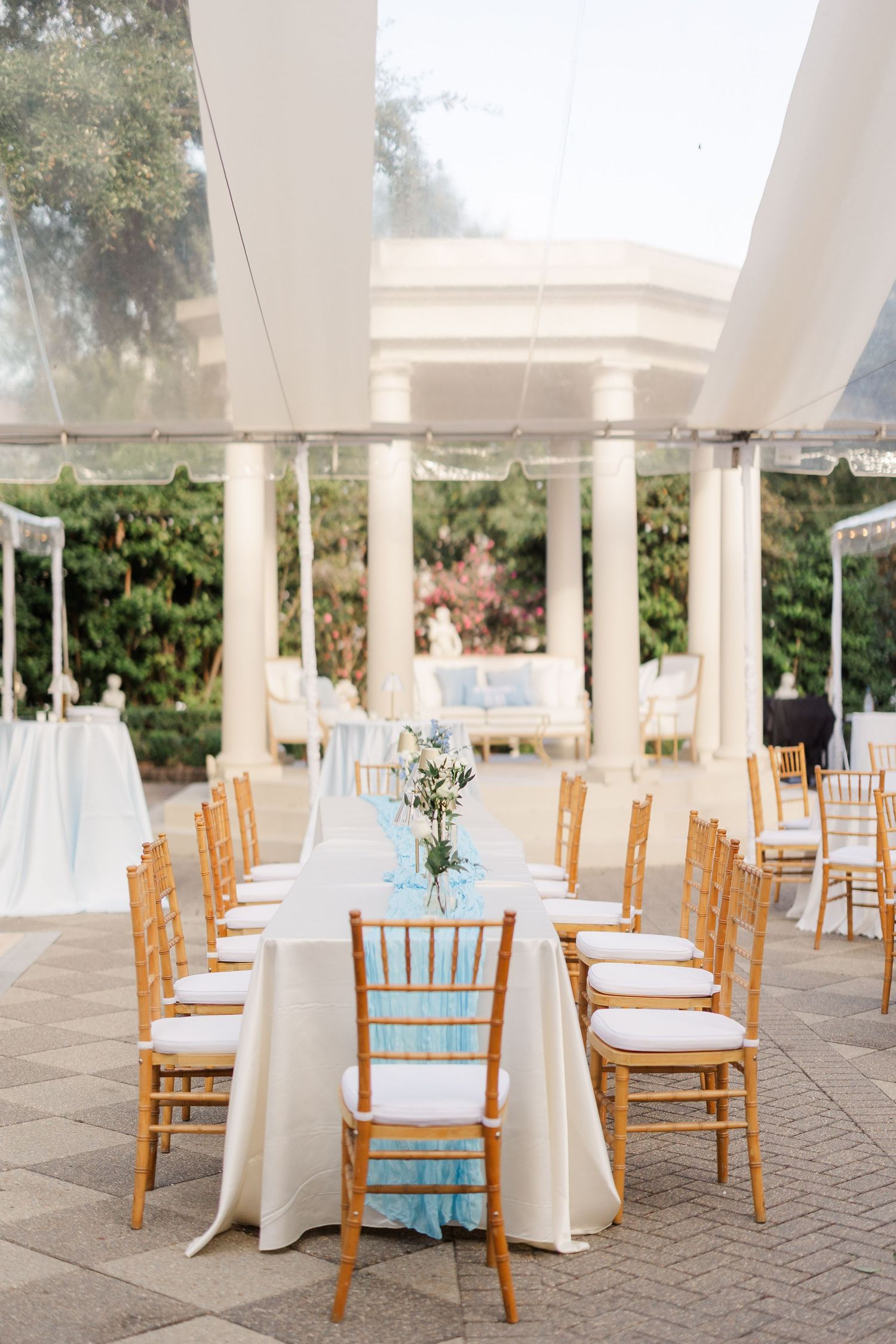 Elegant outdoor wedding reception setup with wooden chairs and white tablecloths under columned pavilion.