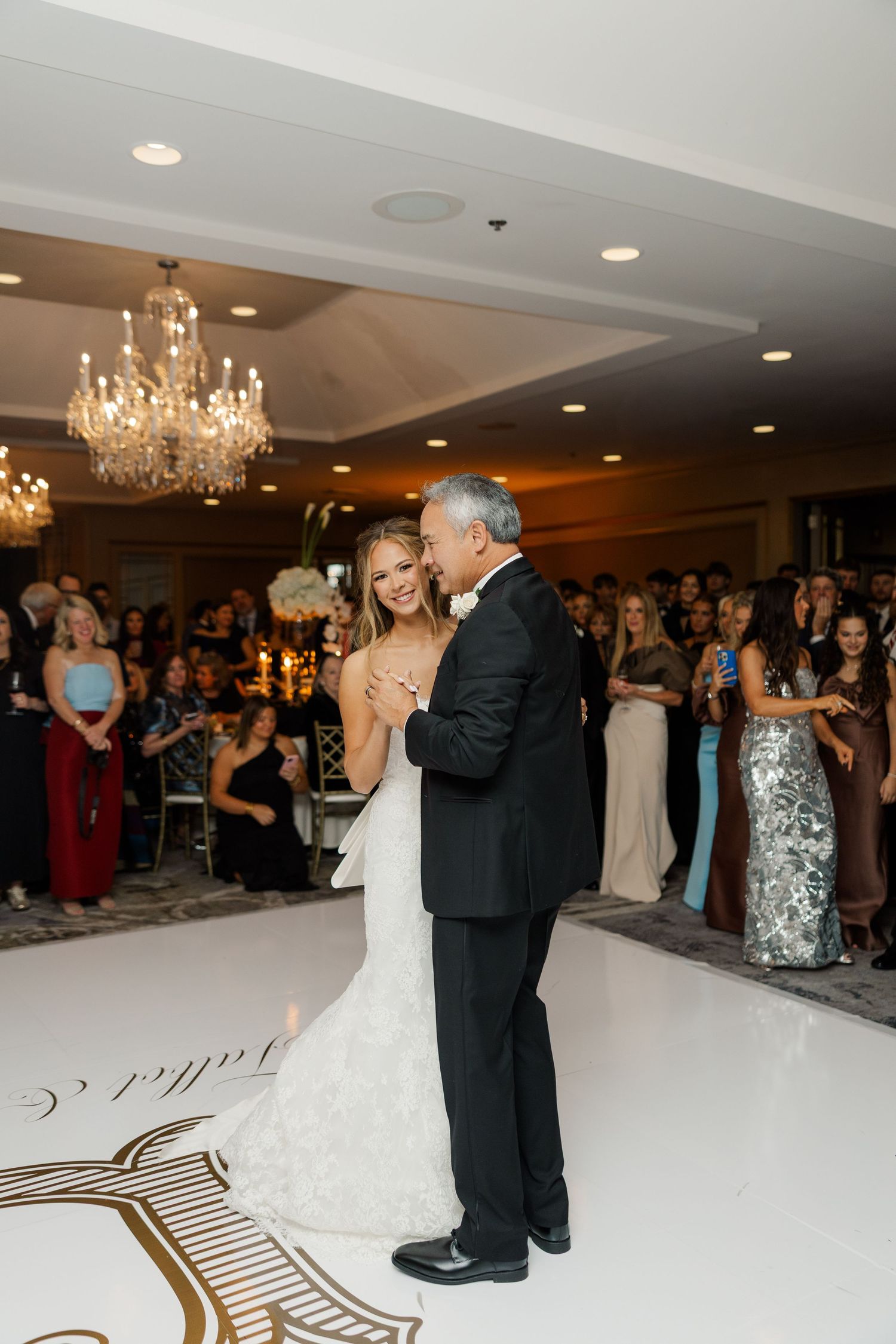 Wedding couple shares first dance on white ballroom floor with swirled gold pattern under crystal chandeliers.