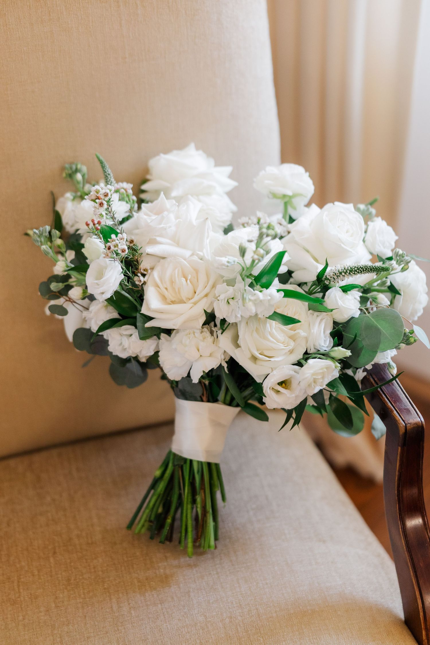 Elegant white rose and greenery wedding bouquet displayed on beige chair.
