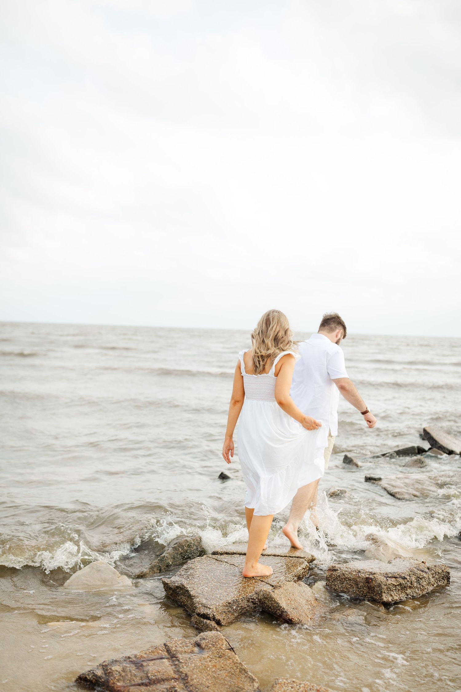A couple walks along rocky coastal shoreline with waves crashing as the woman's white dress flows in the breeze.