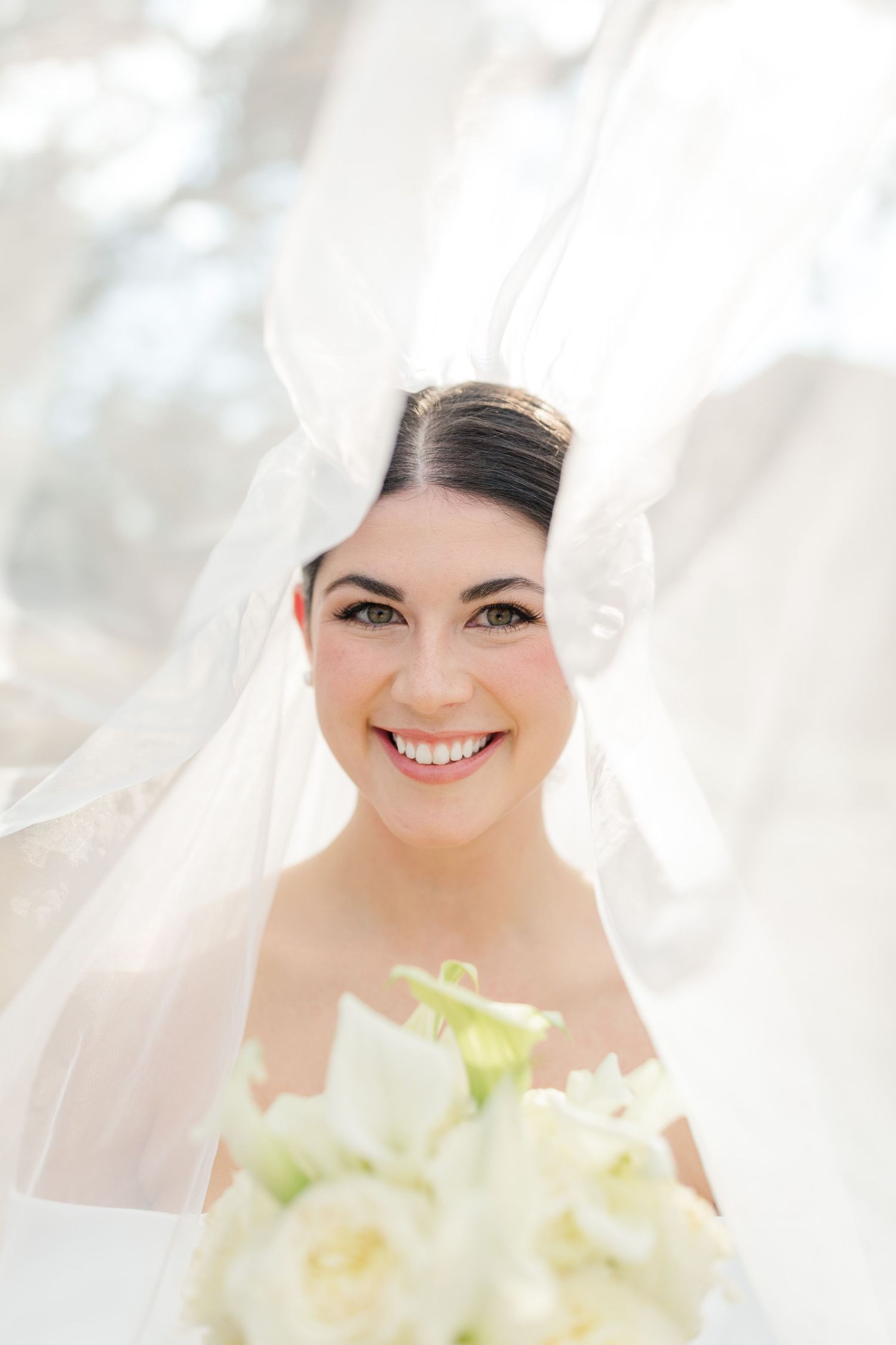 A radiant bride smiles under a flowing white veil while holding a white floral wedding bouquet.