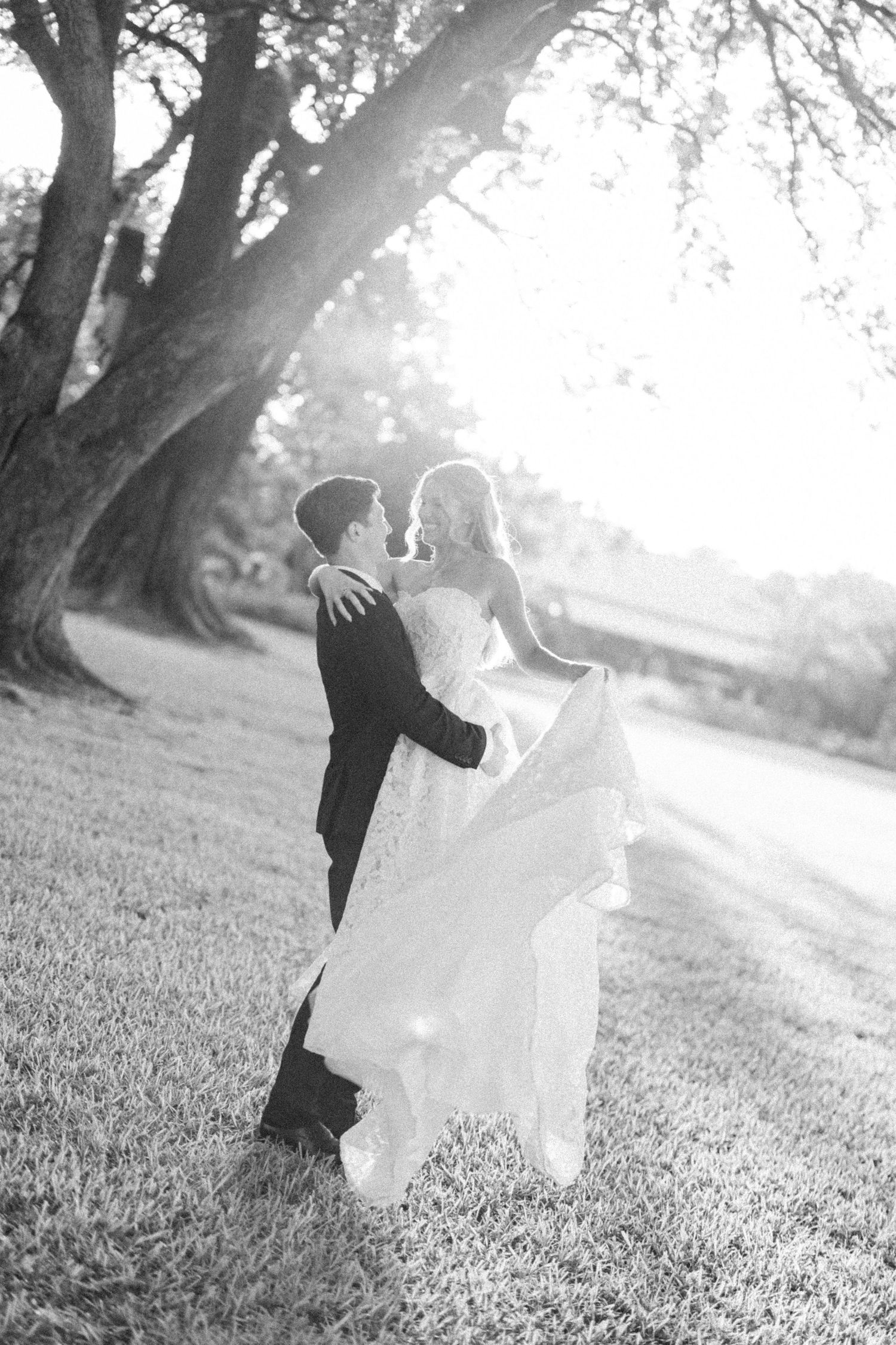 Black and white photo of bride and groom embracing under large oak tree at golden hour.