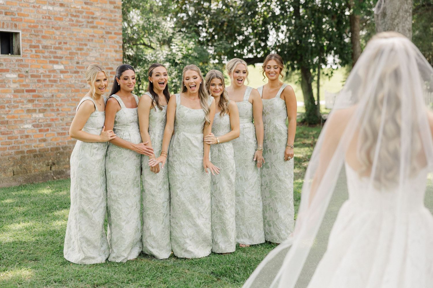 Bride in white gown and veil faces smiling bridesmaids in matching sage green lace dresses outdoors near brick building.