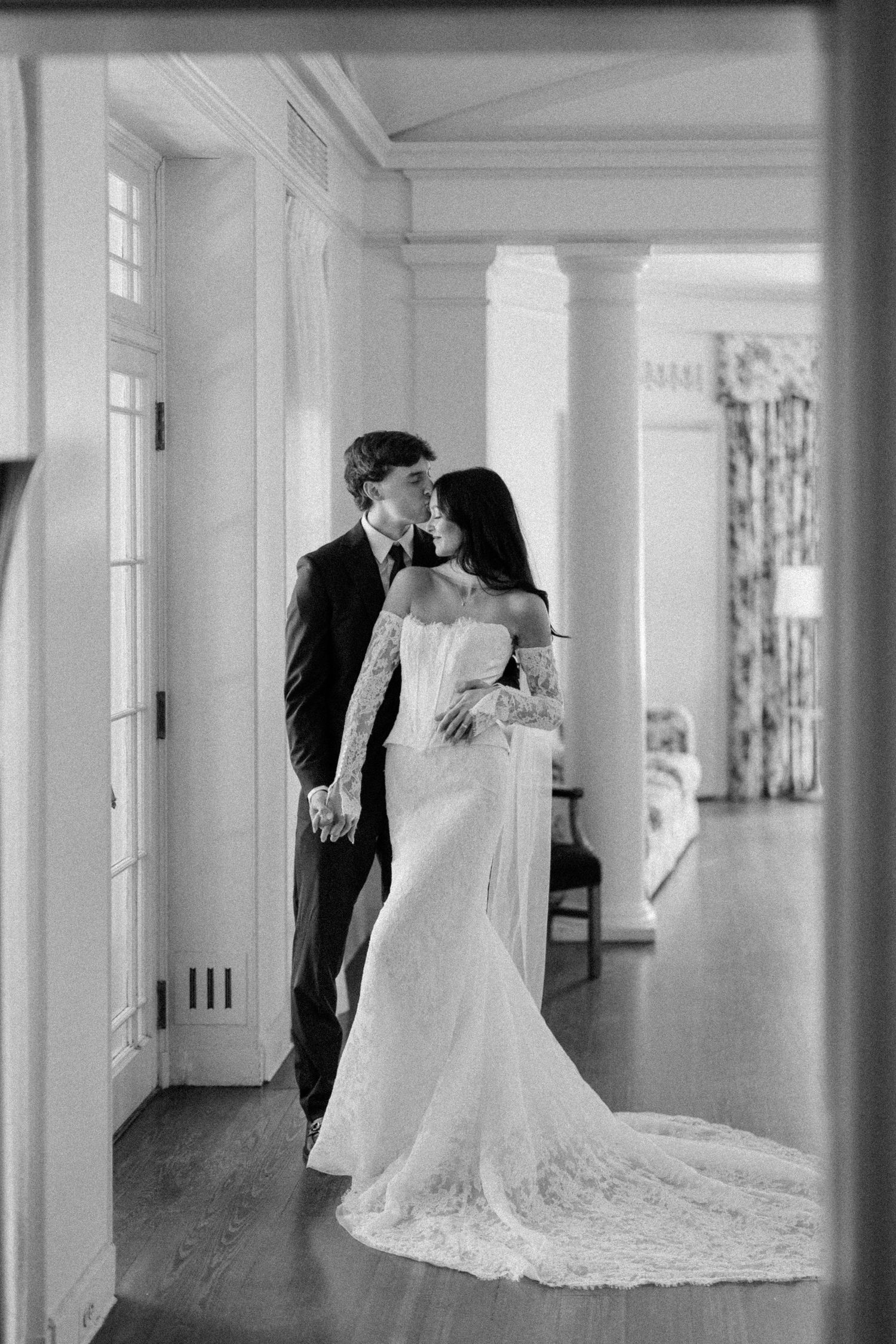 Black and white photo of a bride and groom sharing a tender kiss in an elegant hallway with white columns.