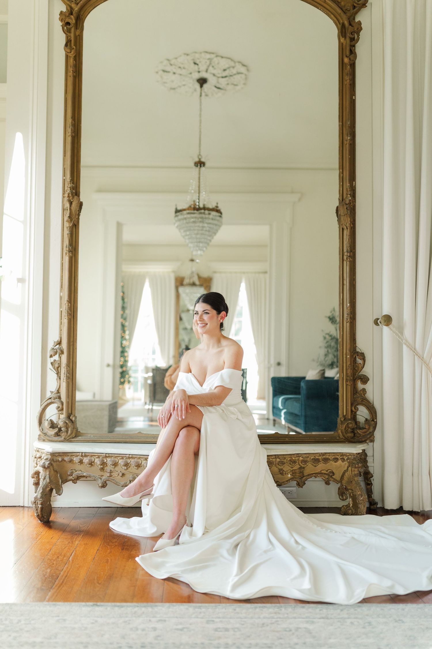 Elegant bridal portrait reflected in an ornate gold mirror showing a flowing white gown and hardwood floors.