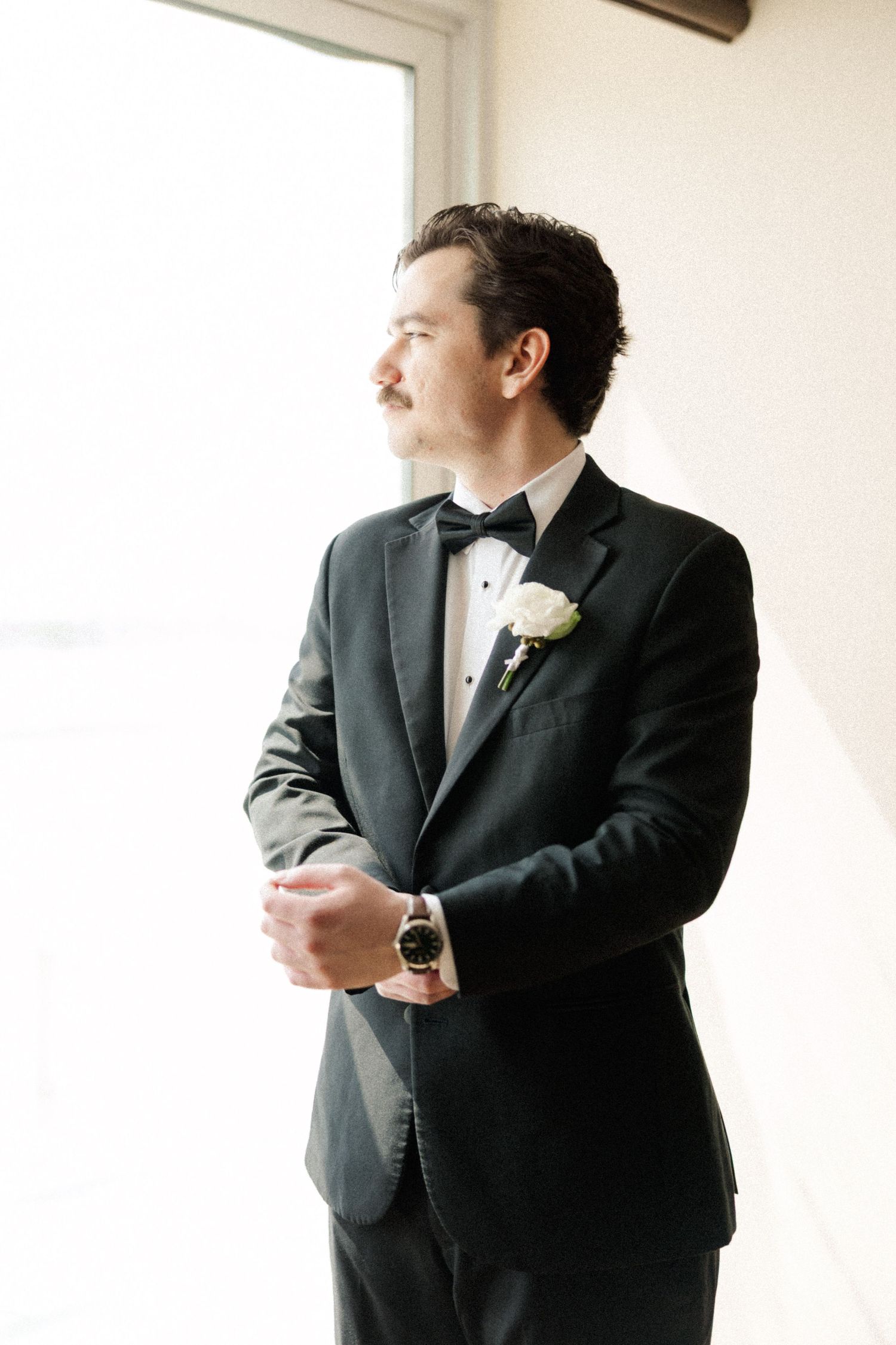 Dapper groom in black tuxedo with bow tie and white boutonniere gazes out window on wedding day.
