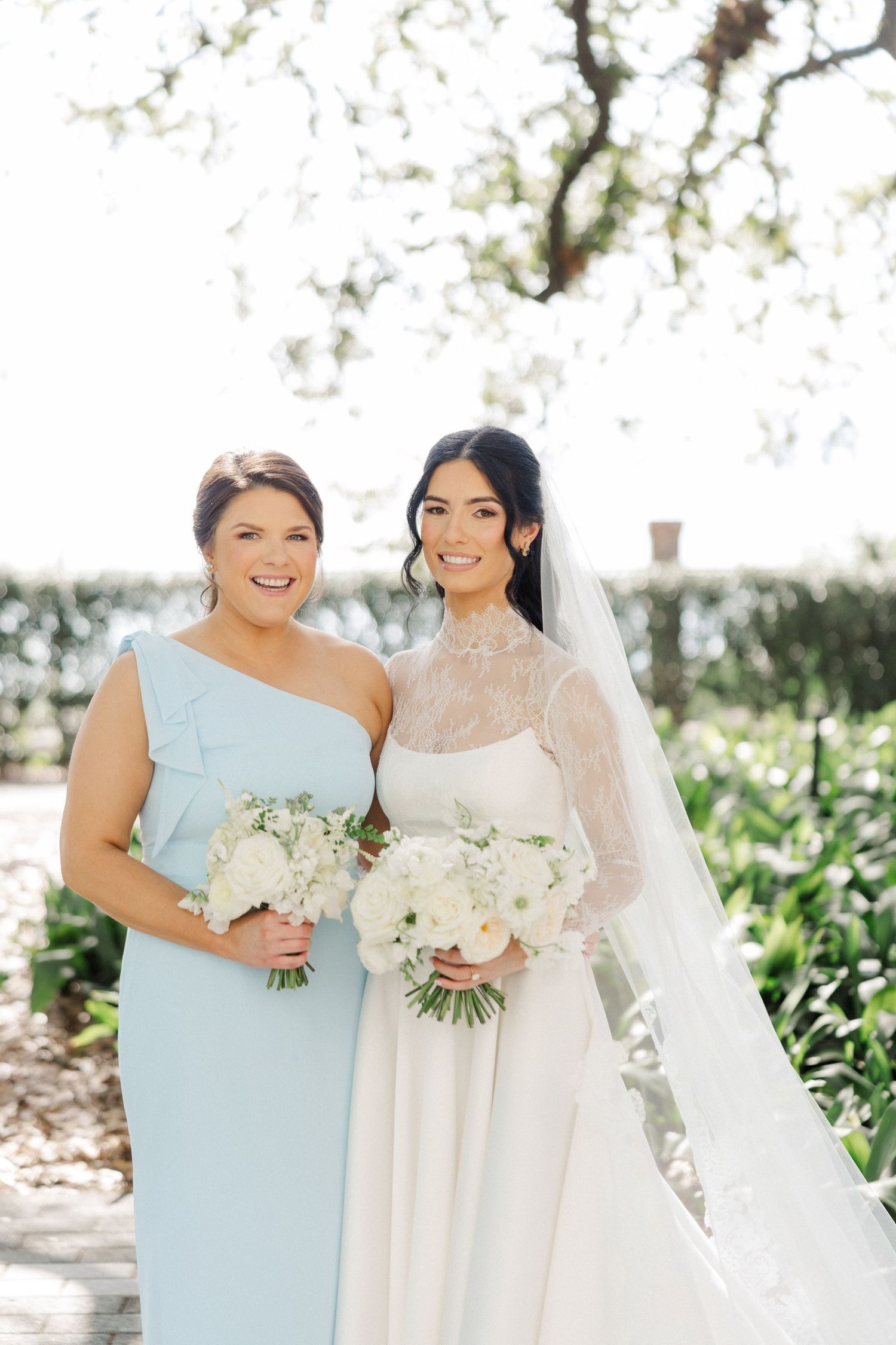 Bride in white lace gown with veil and bridesmaid in light blue one-shoulder dress holding white bouquets outdoors.