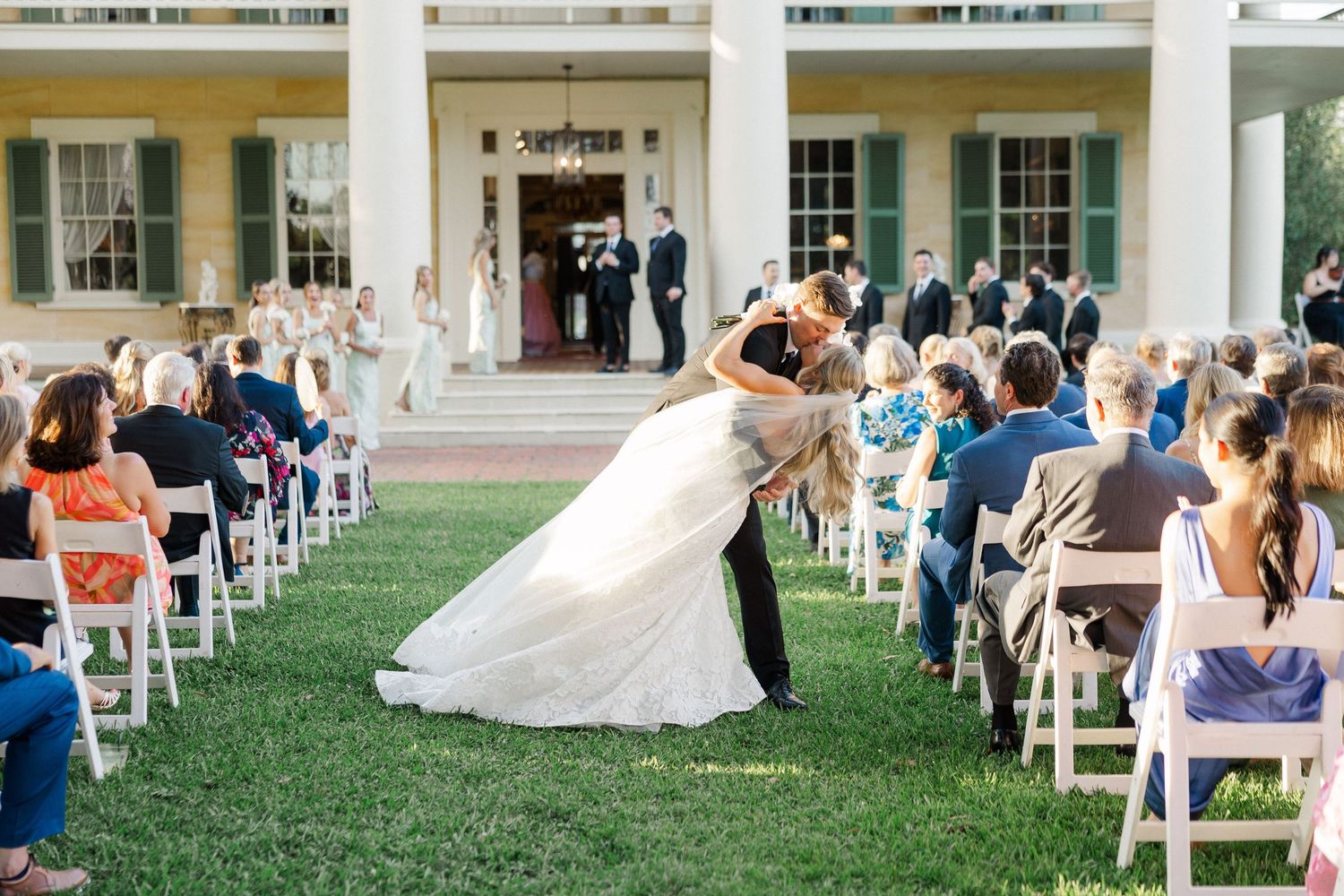 Groom dips bride for a kiss at outdoor wedding ceremony on lawn in front of a white columned estate.