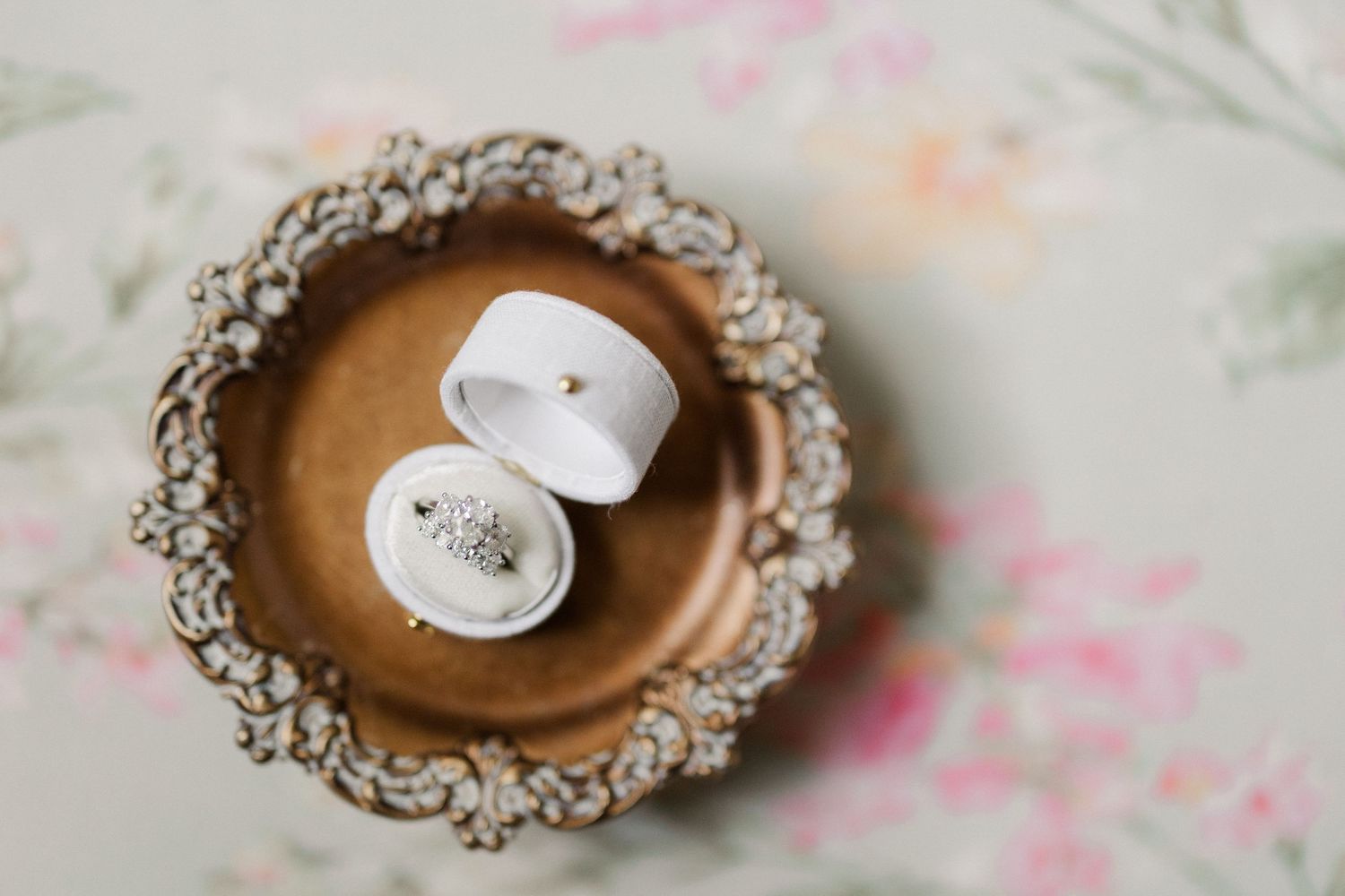 Wedding rings in a white velvet box displayed on an ornate vintage gold trinket dish with floral background.