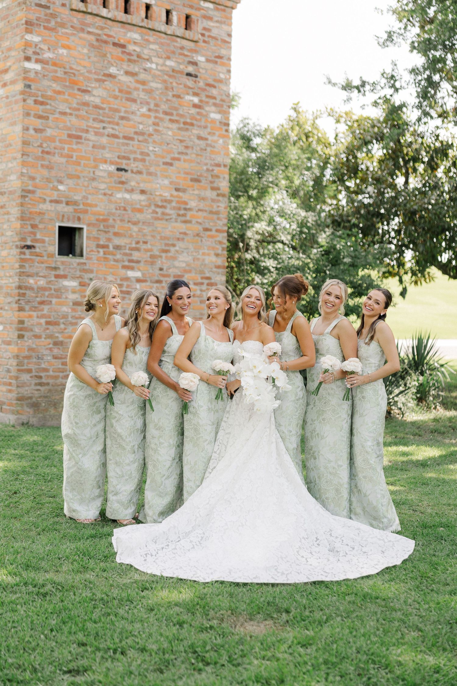 Smiling bride in lace wedding gown laughs with bridesmaids in sage green dresses beside rustic brick tower.