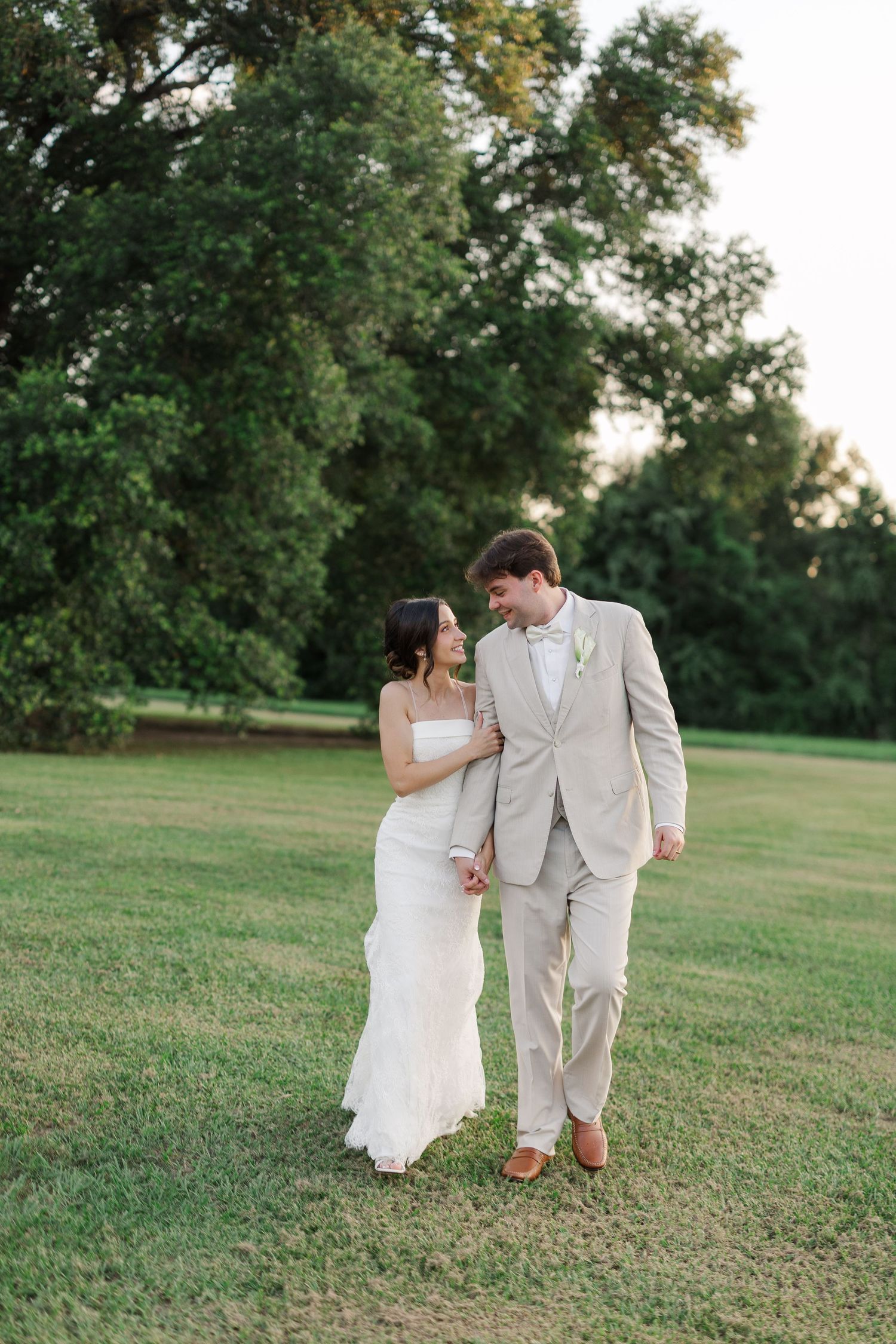 Bride and groom walking hand in hand across green lawn at golden hour near lush trees.