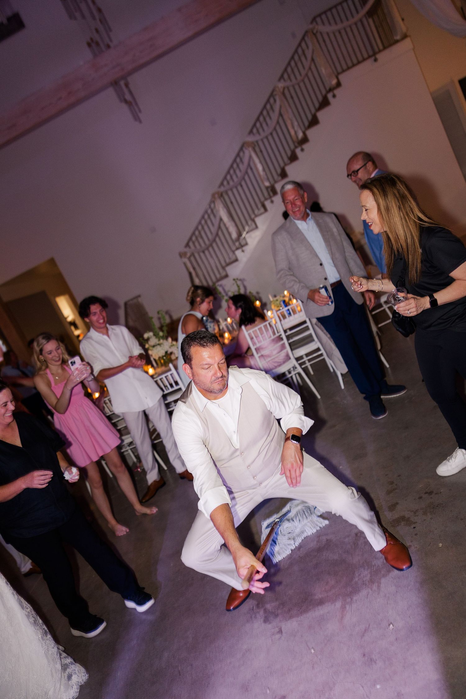 Groom in white suit energetically dancing low on reception floor surrounded by cheering guests.