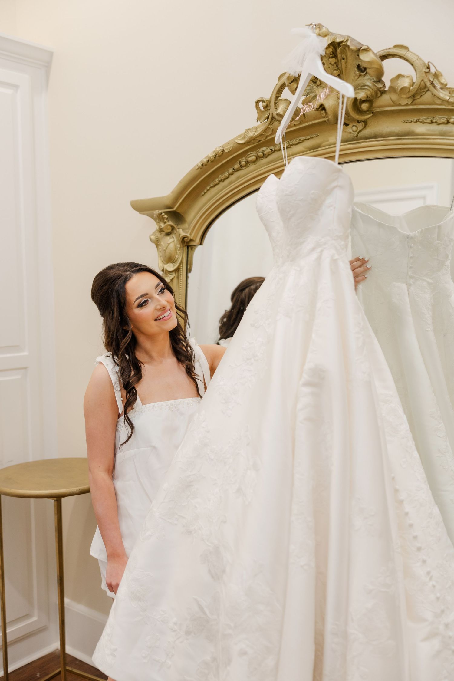 Beautiful white wedding dress displayed in an elegant room with ornate gold mirror and white walls.