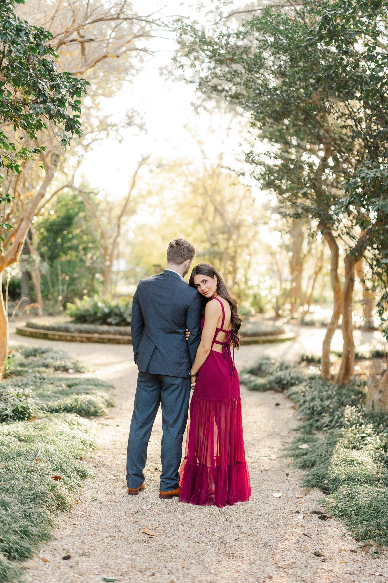 A couple walks together on a wooded path, with the woman wearing a burgundy dress and man in navy suit.