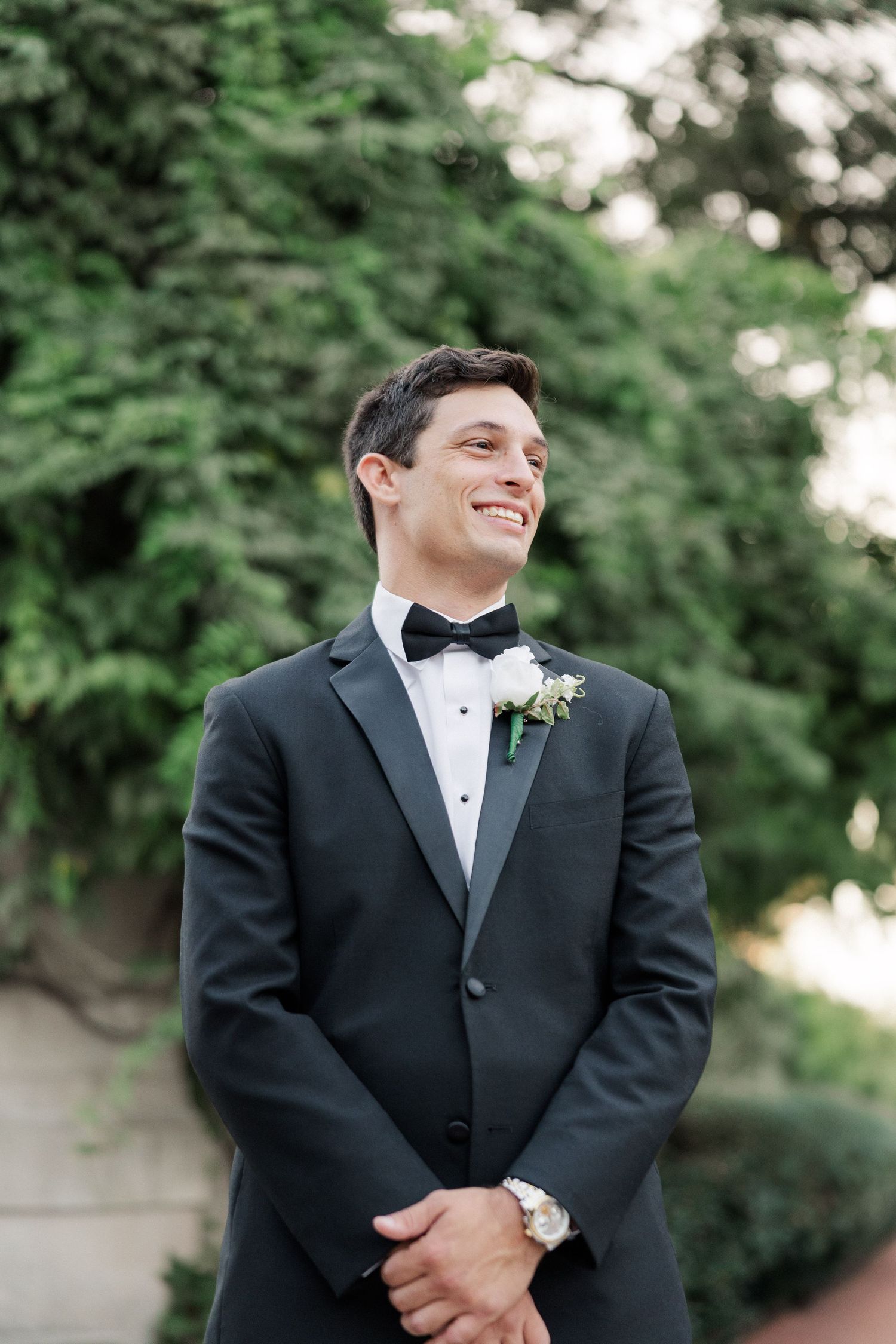 Groom in classic black tuxedo smiles while wearing boutonniere outdoors.